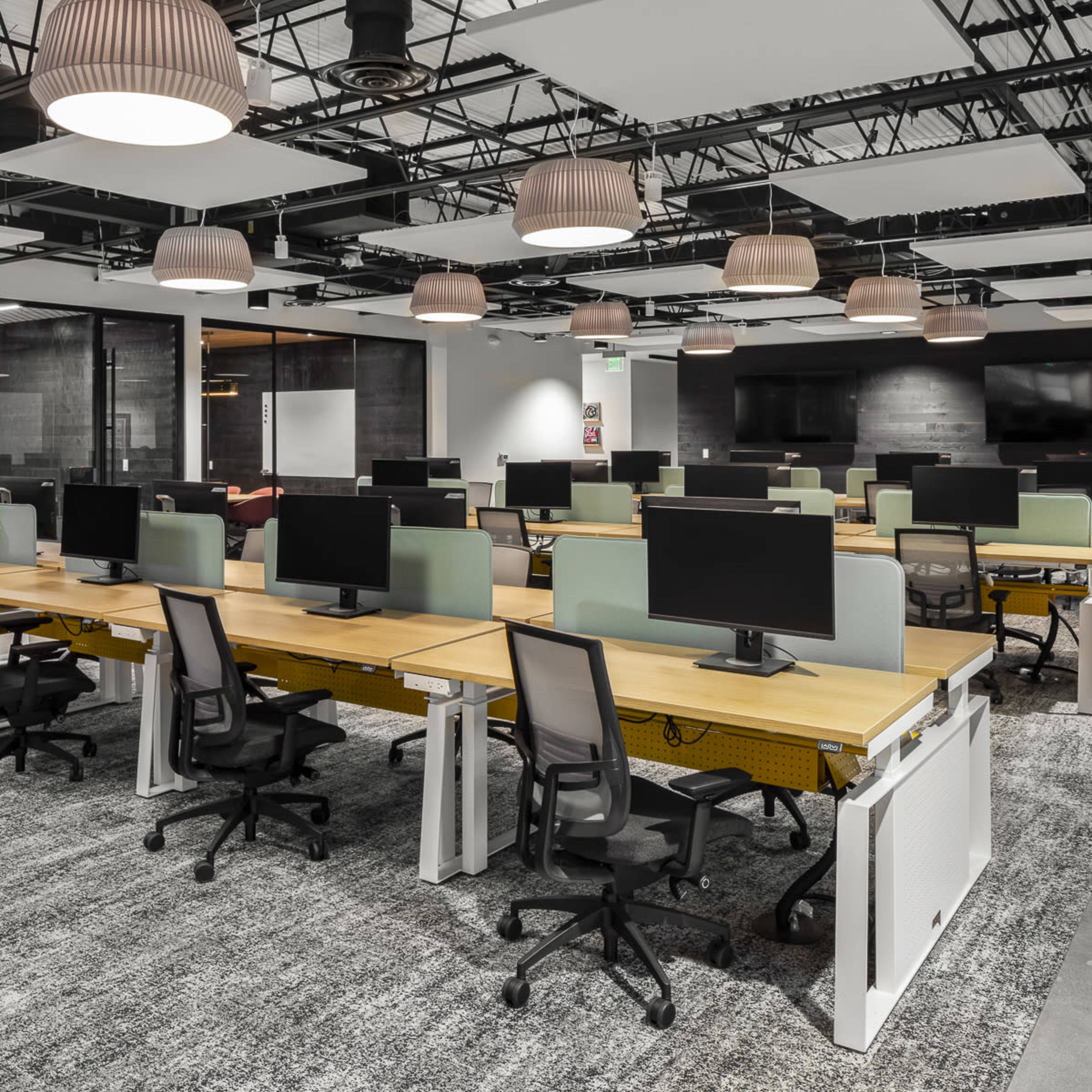 A modern office space with rows of workstations, each equipped with computers and ergonomic chairs, under hanging light fixtures.