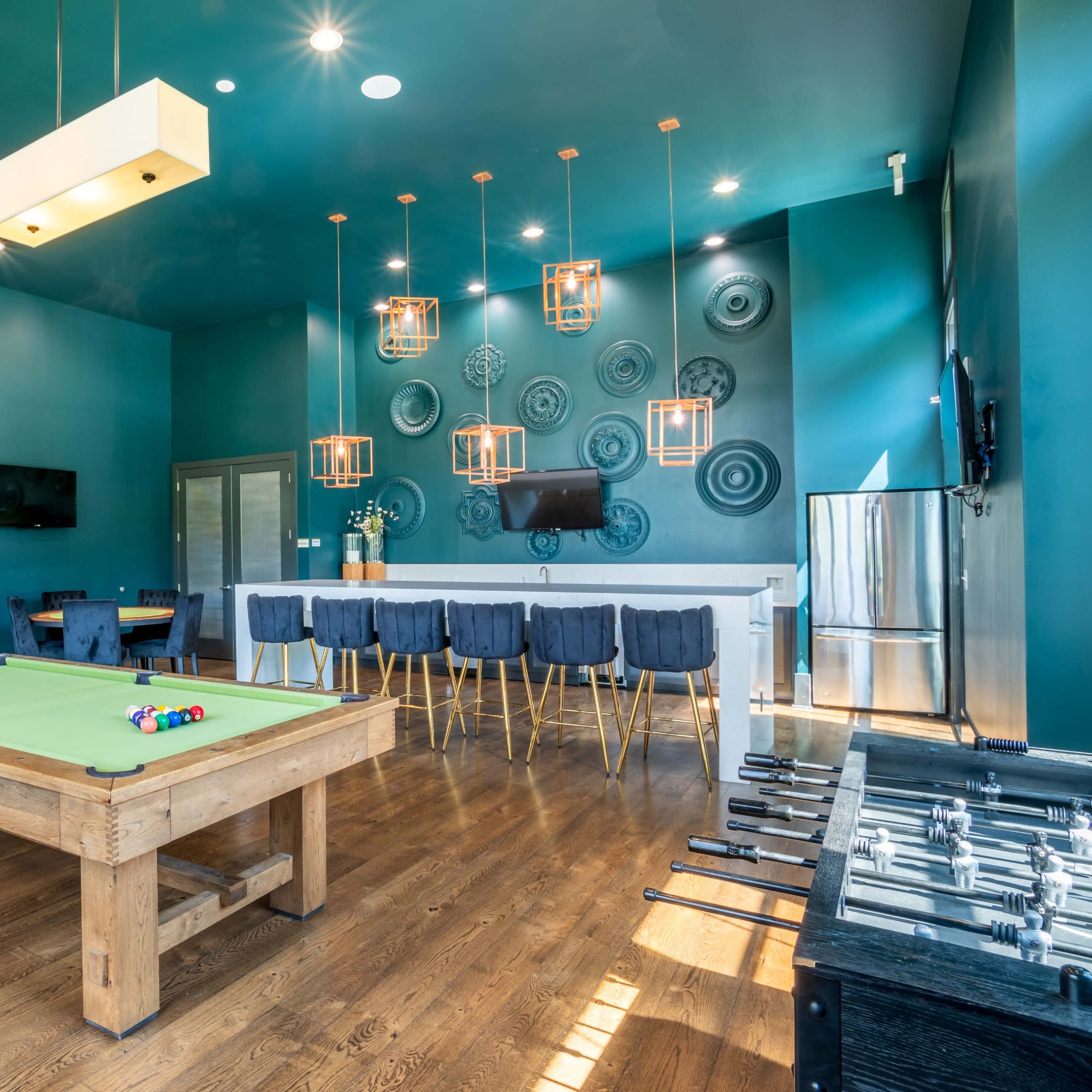 A game room featuring a pool table, a foosball table, a bar area with high stools, and various decorative elements on the walls.