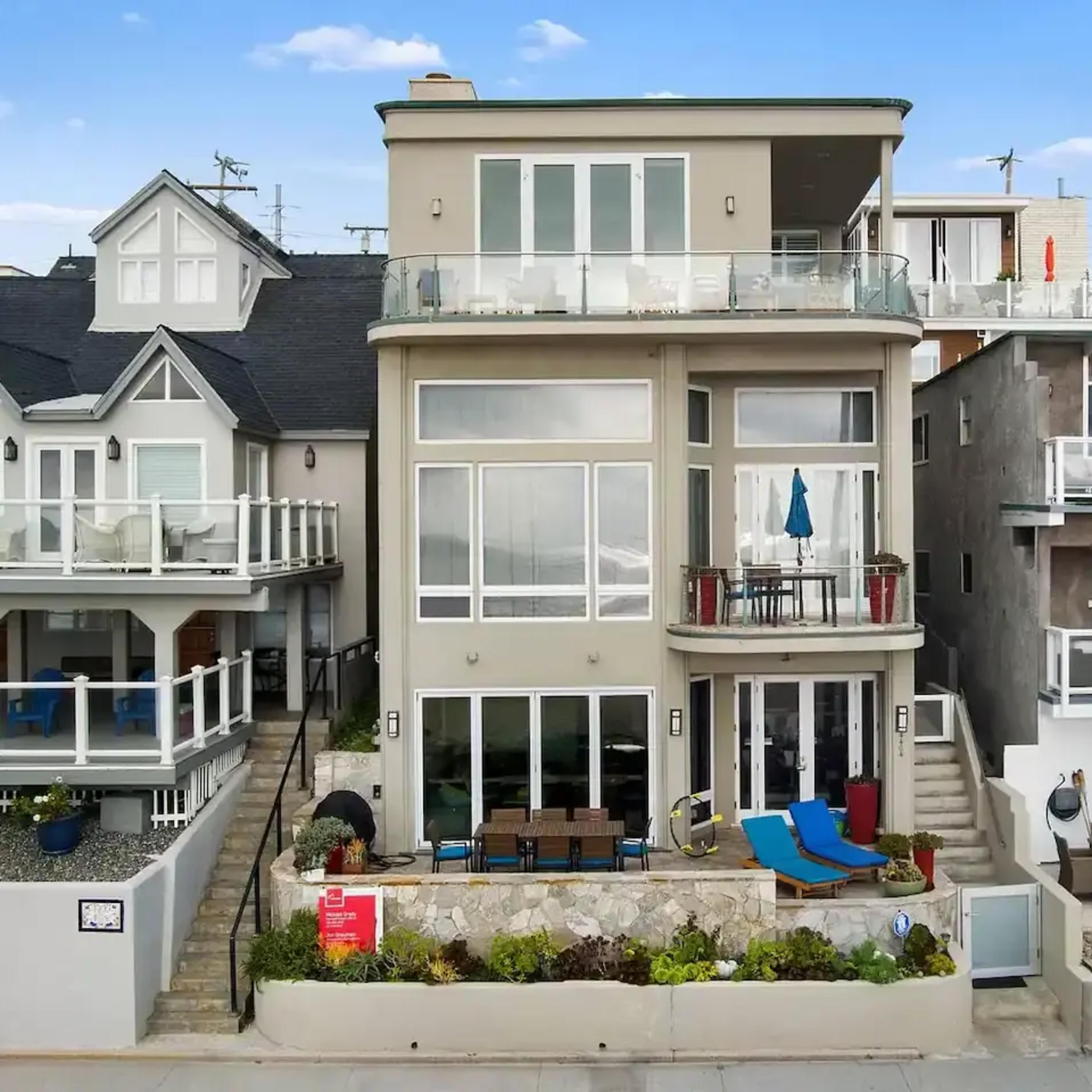 A modern beach house with large glass windows and multiple outdoor seating areas, situated adjacent to other similar homes along a coastal street.