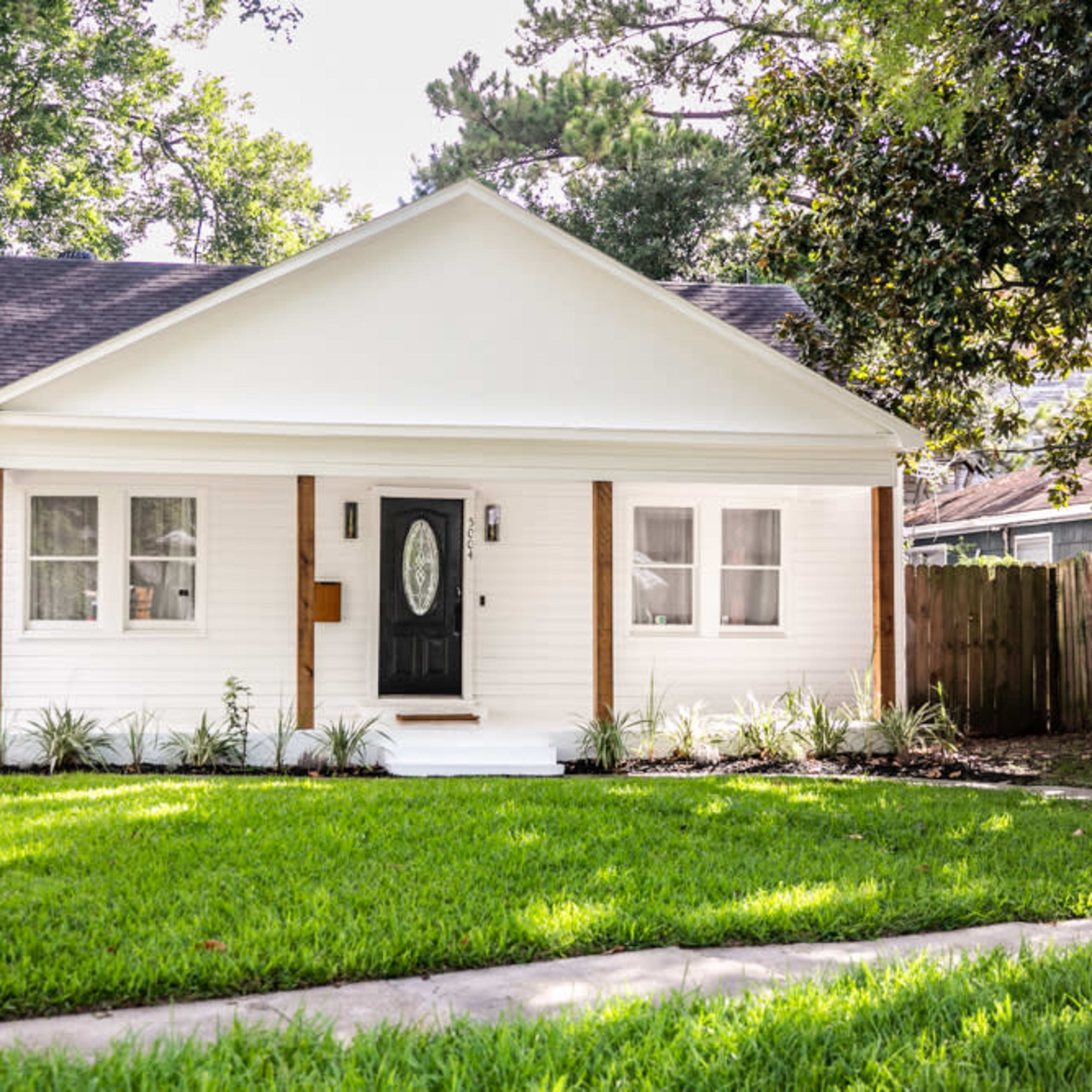 A single-story white house with a black front door is surrounded by a green lawn and trees.