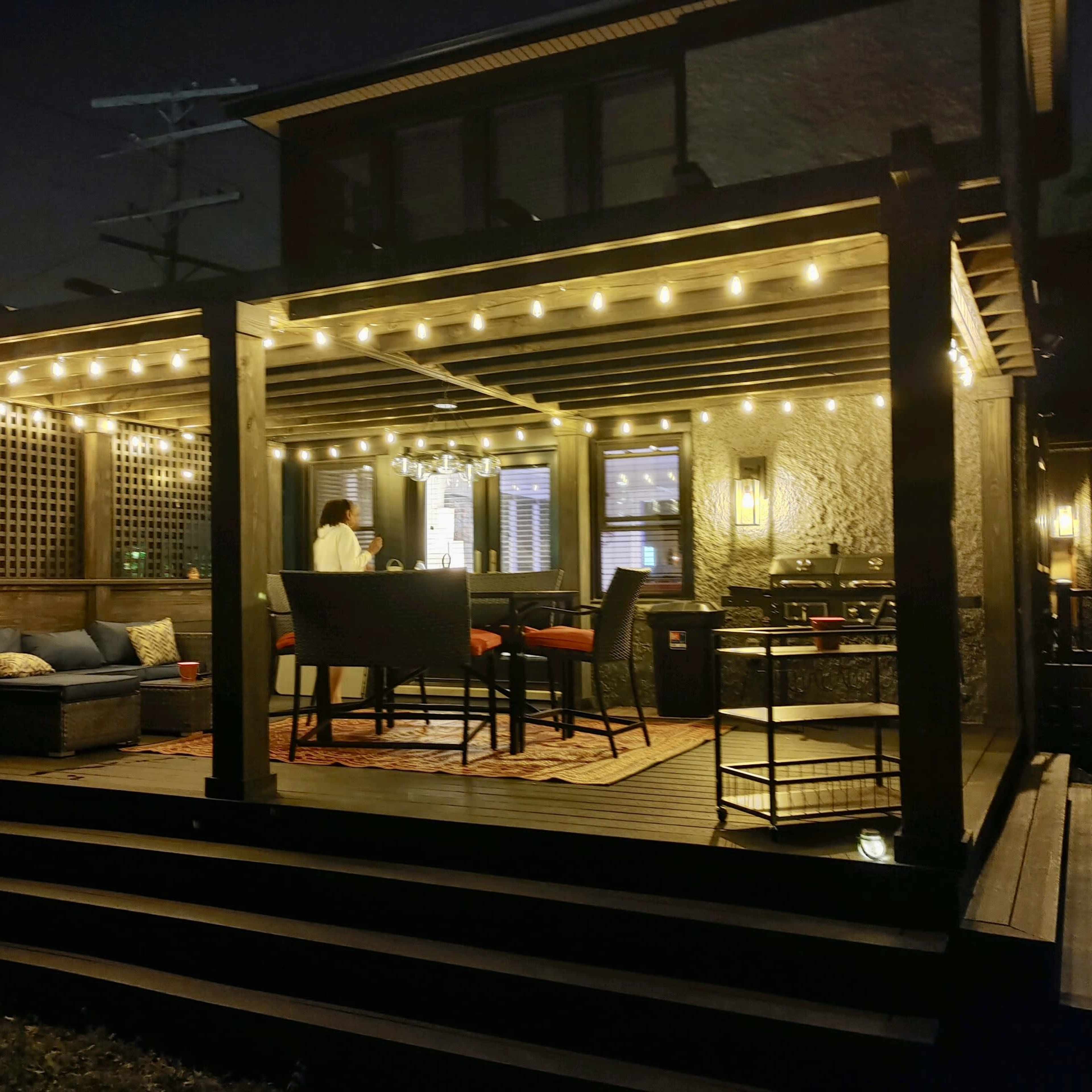 A well-lit outdoor patio features a table and chairs beneath a covered pergola, with string lights illuminating the space at night.