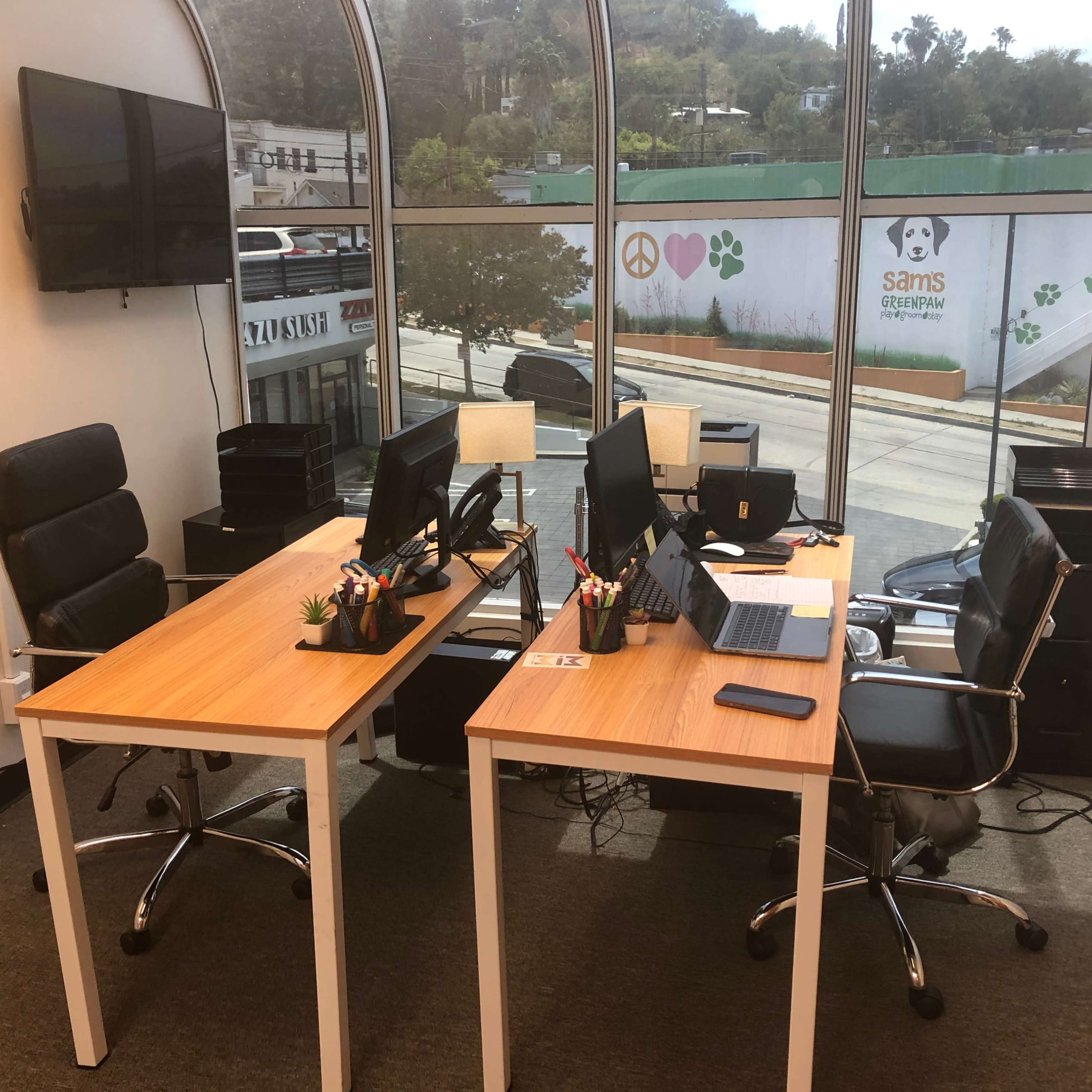 A two-desk office setup with computers and office supplies, located near large windows overlooking a street.