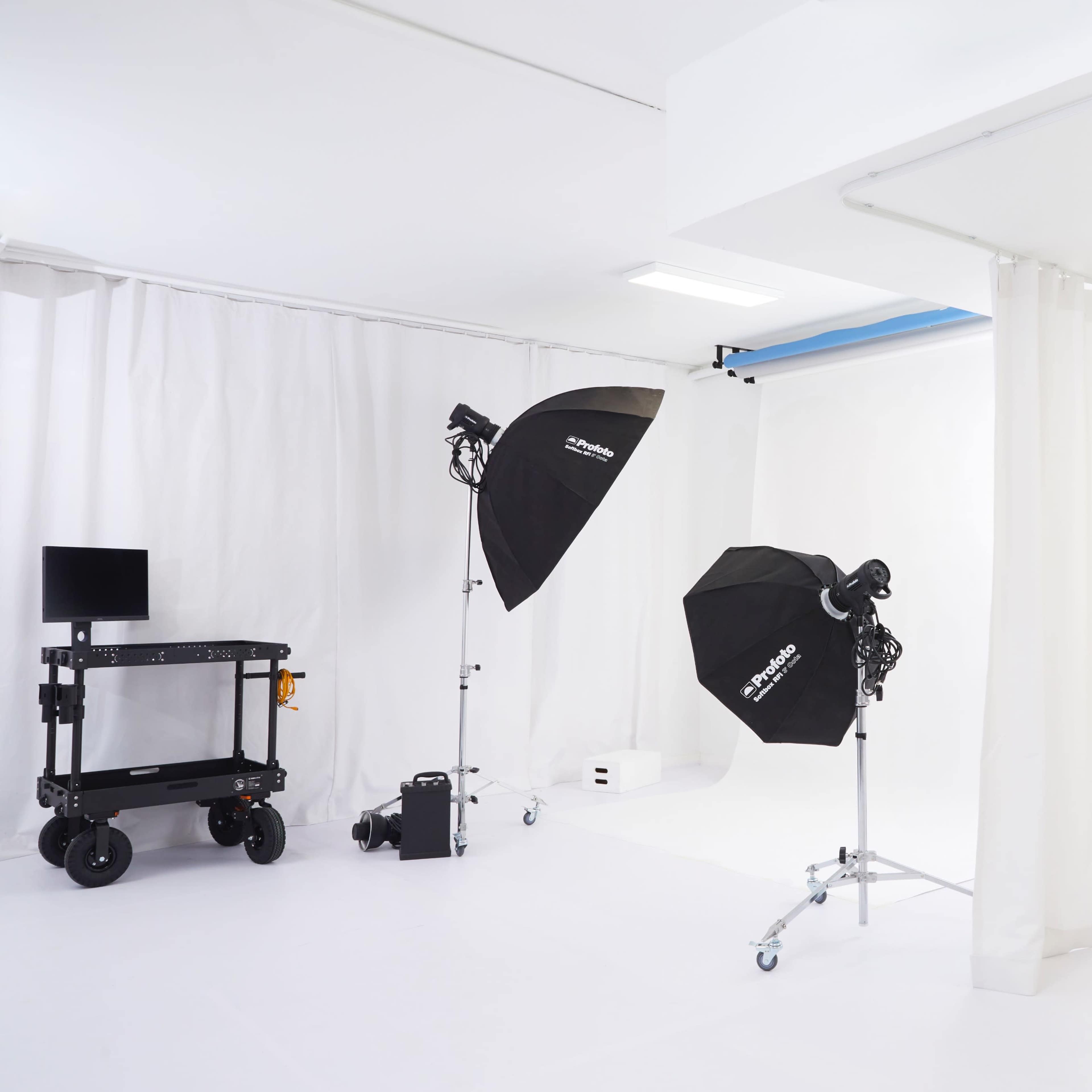 A photography studio features white walls, lighting equipment on stands, a portable cart, and a large mirror surrounded by curtains.