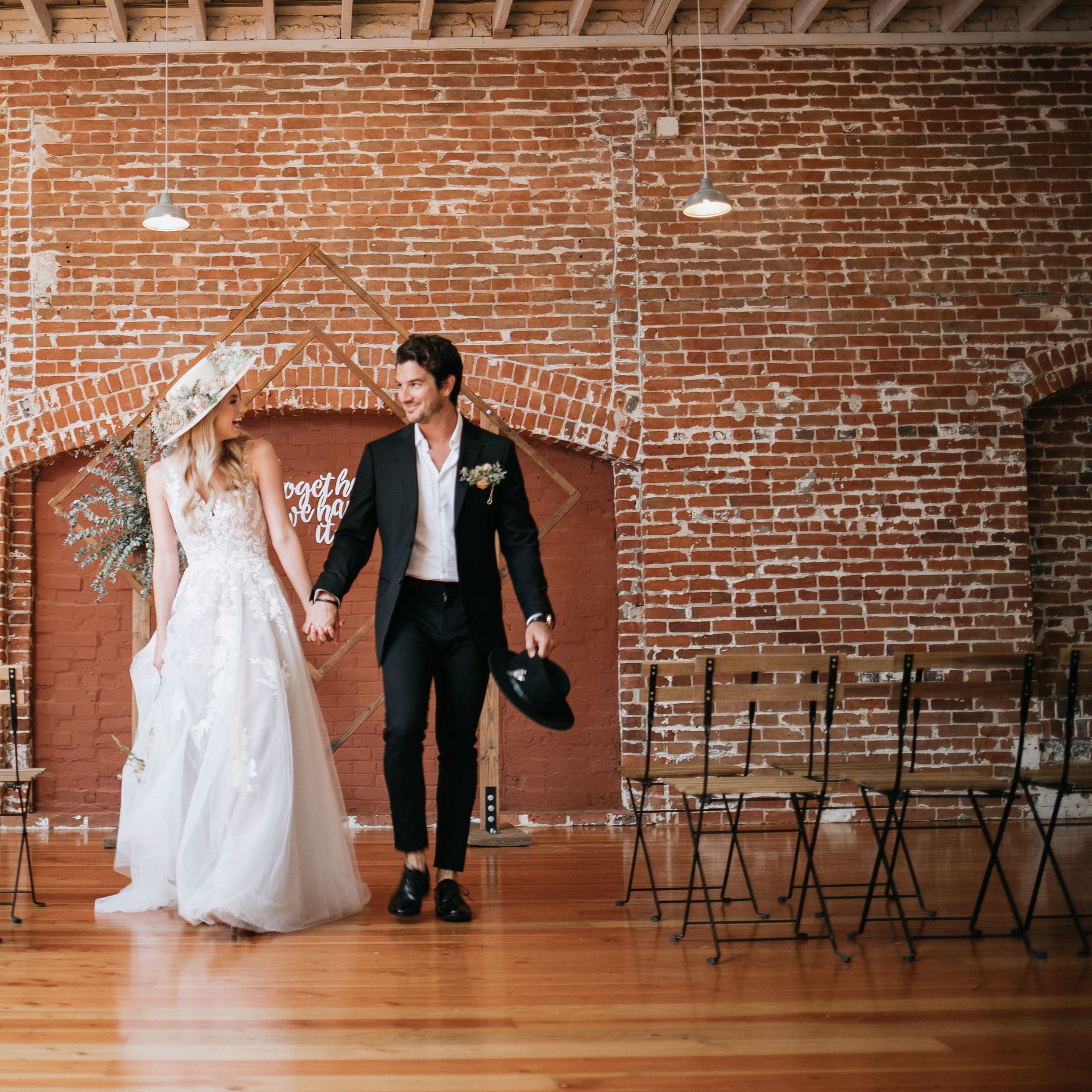 A couple in formal wedding attire walks hand in hand in front of a brick wall adorned with decorative elements, with metal chairs arranged for a ceremony.