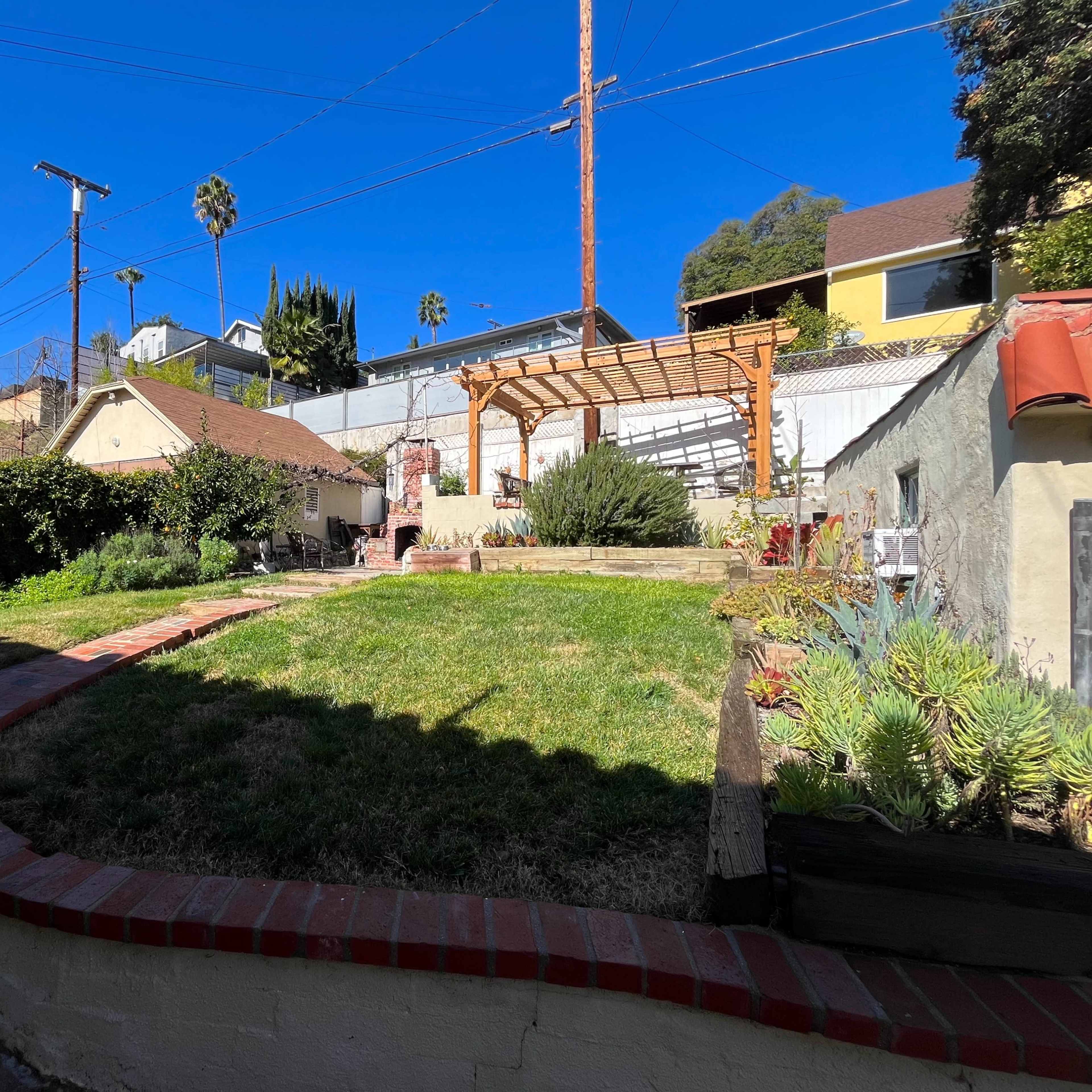 A backyard with a grass lawn, a wooden pergola, and various plants, surrounded by residential buildings and a clear blue sky.