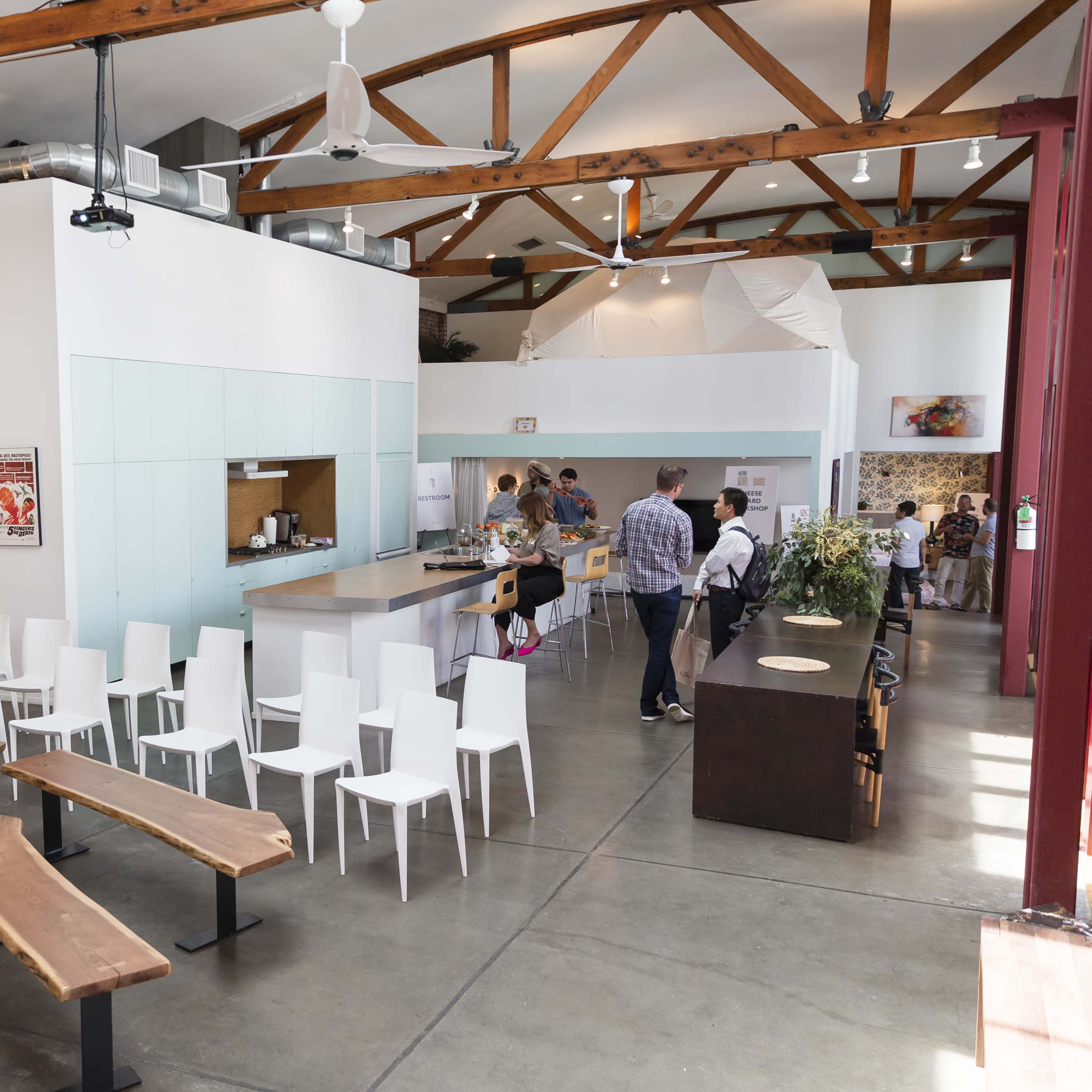 A spacious, modern café interior with wooden beams, a seating area with benches, a service counter, and patrons engaged in activities around the space.