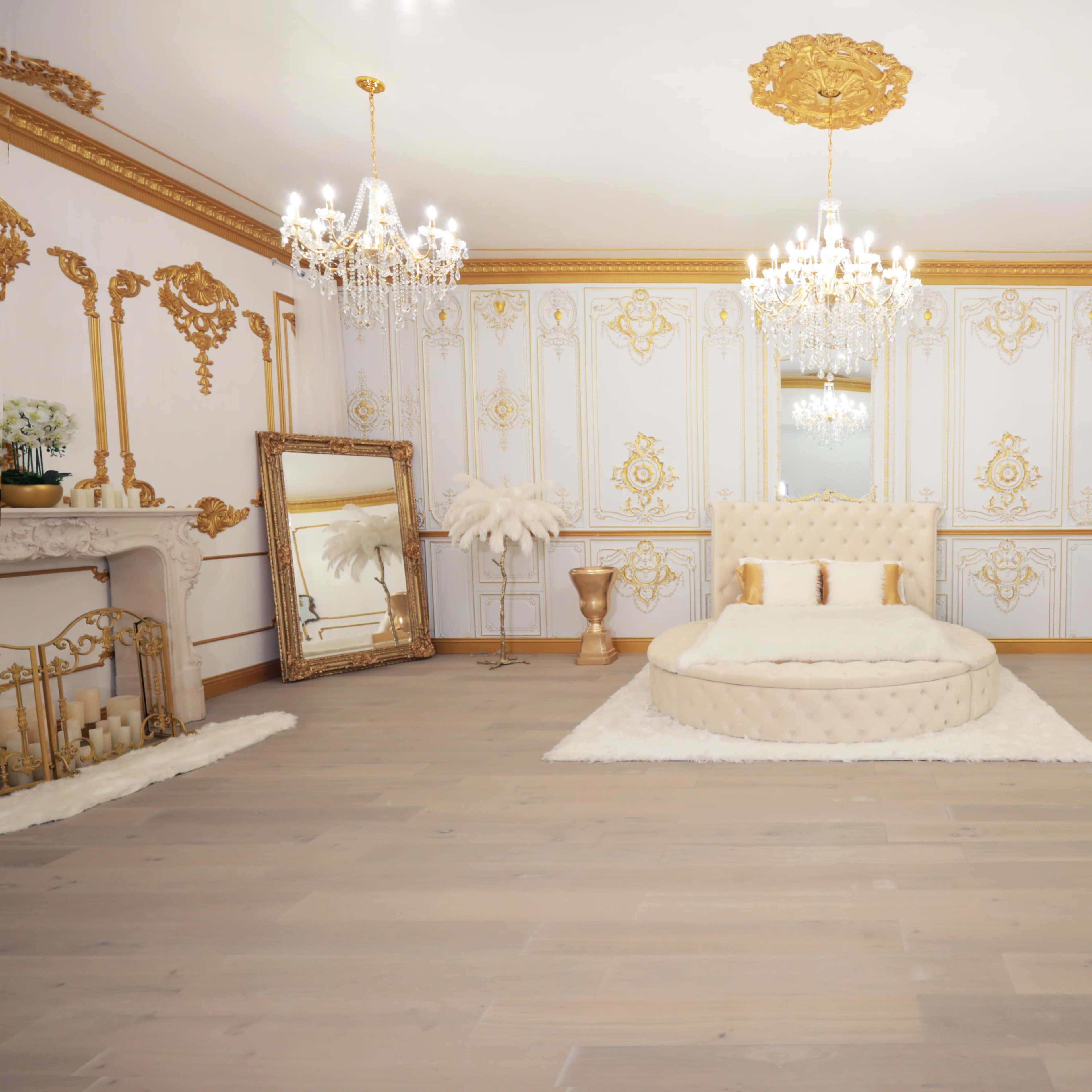 An opulent bedroom featuring a circular tufted bed, ornate chandeliers, and decorative wall panels with gold accents.