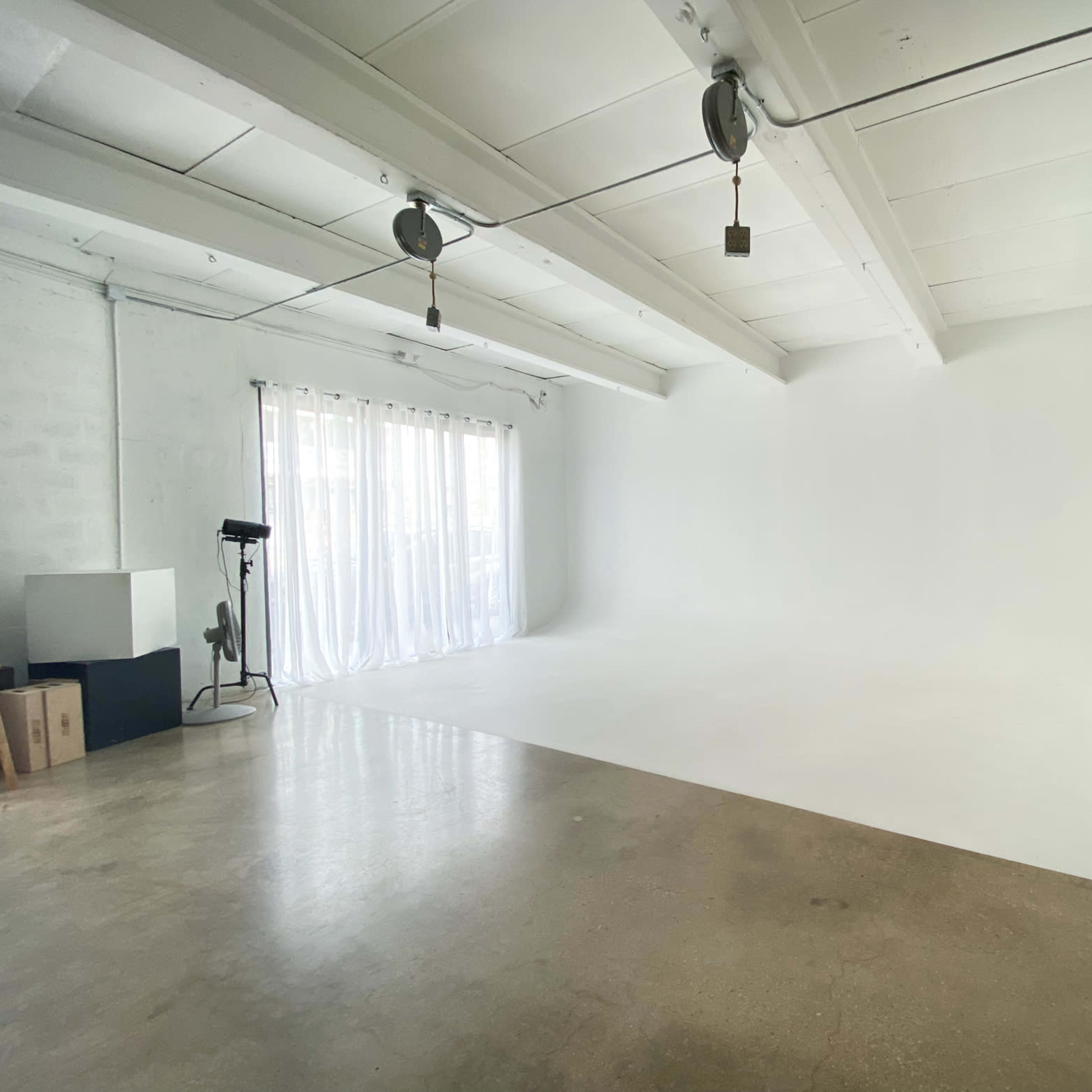 An empty studio space with white walls, a concrete floor, and large windows featuring sheer curtains.