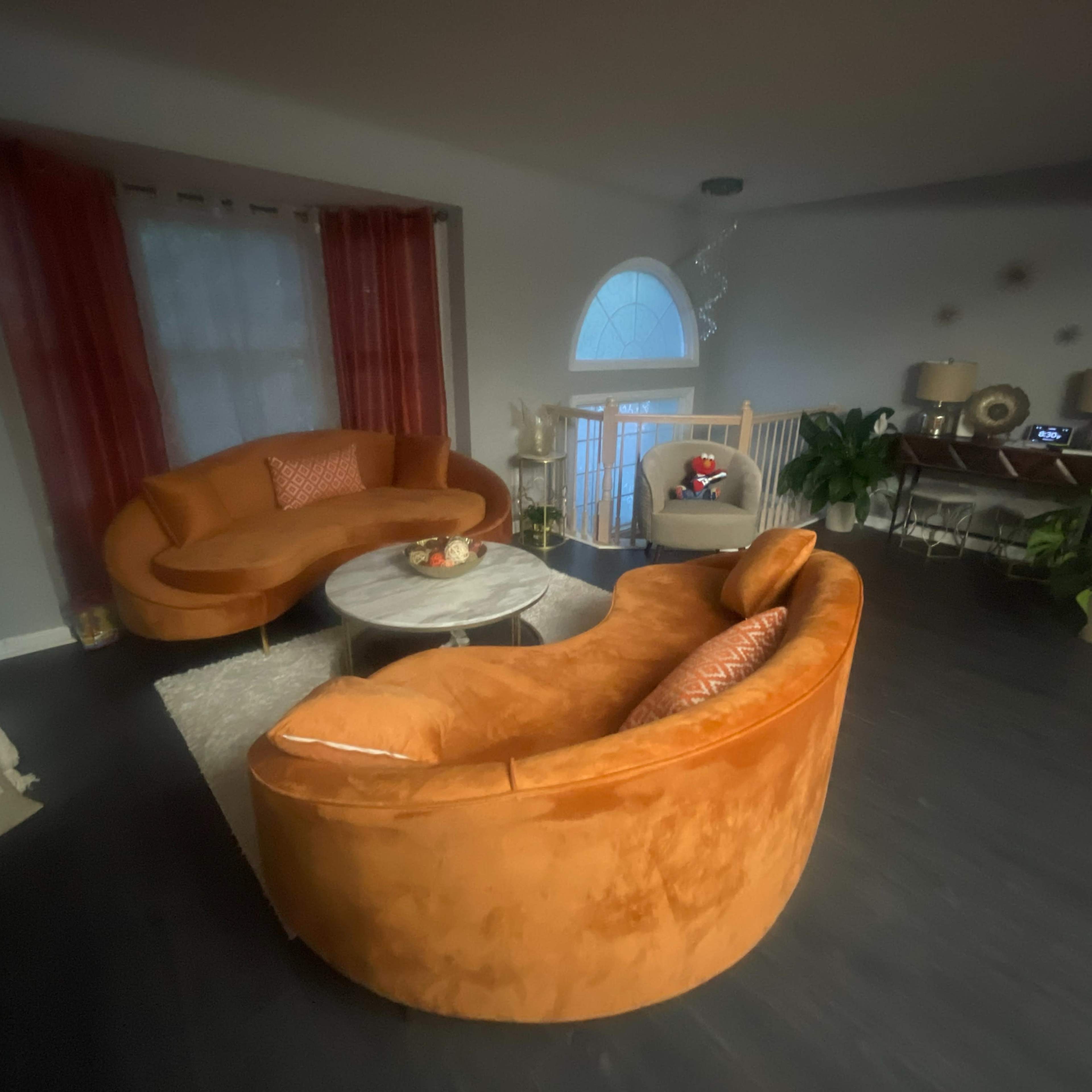 A living room featuring two orange sofas around a circular coffee table, with a light-colored rug and a staircase in the background.