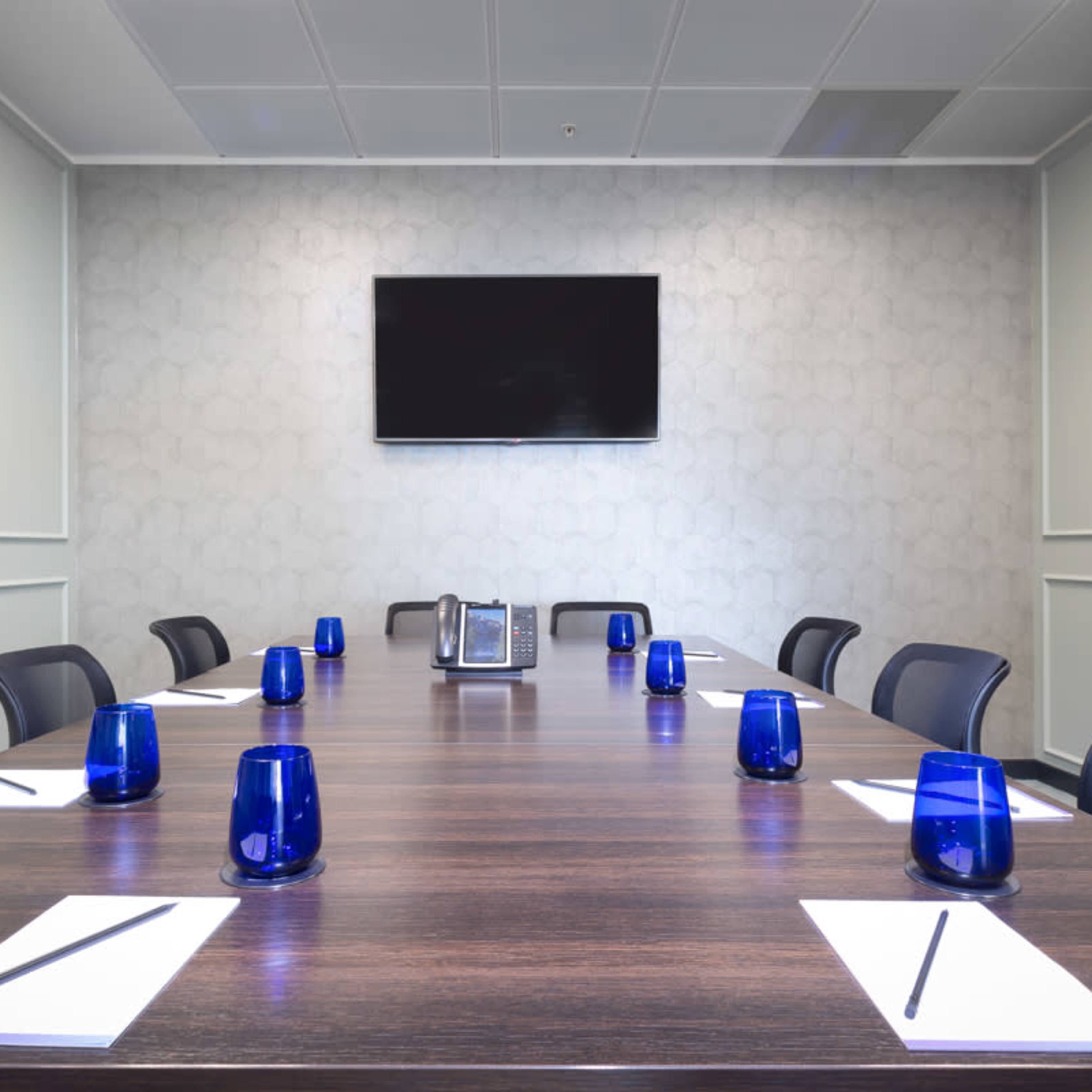 A modern conference room features a long wooden table set with blue glass candle holders, notepads, and chairs arranged around it, with a television mounted on the wall.