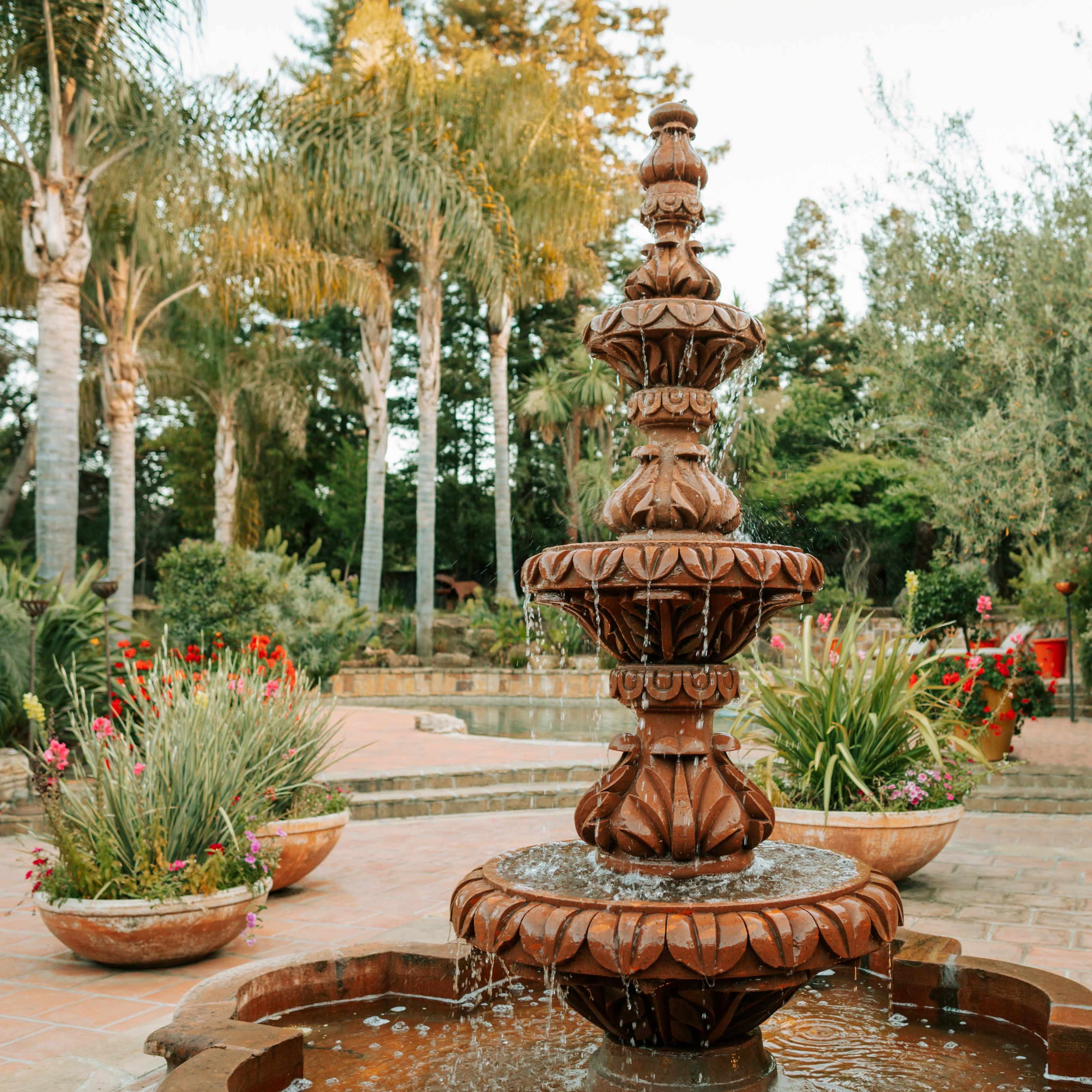 A tiered stone fountain sits in the center of a landscaped garden, surrounded by potted plants and palm trees.