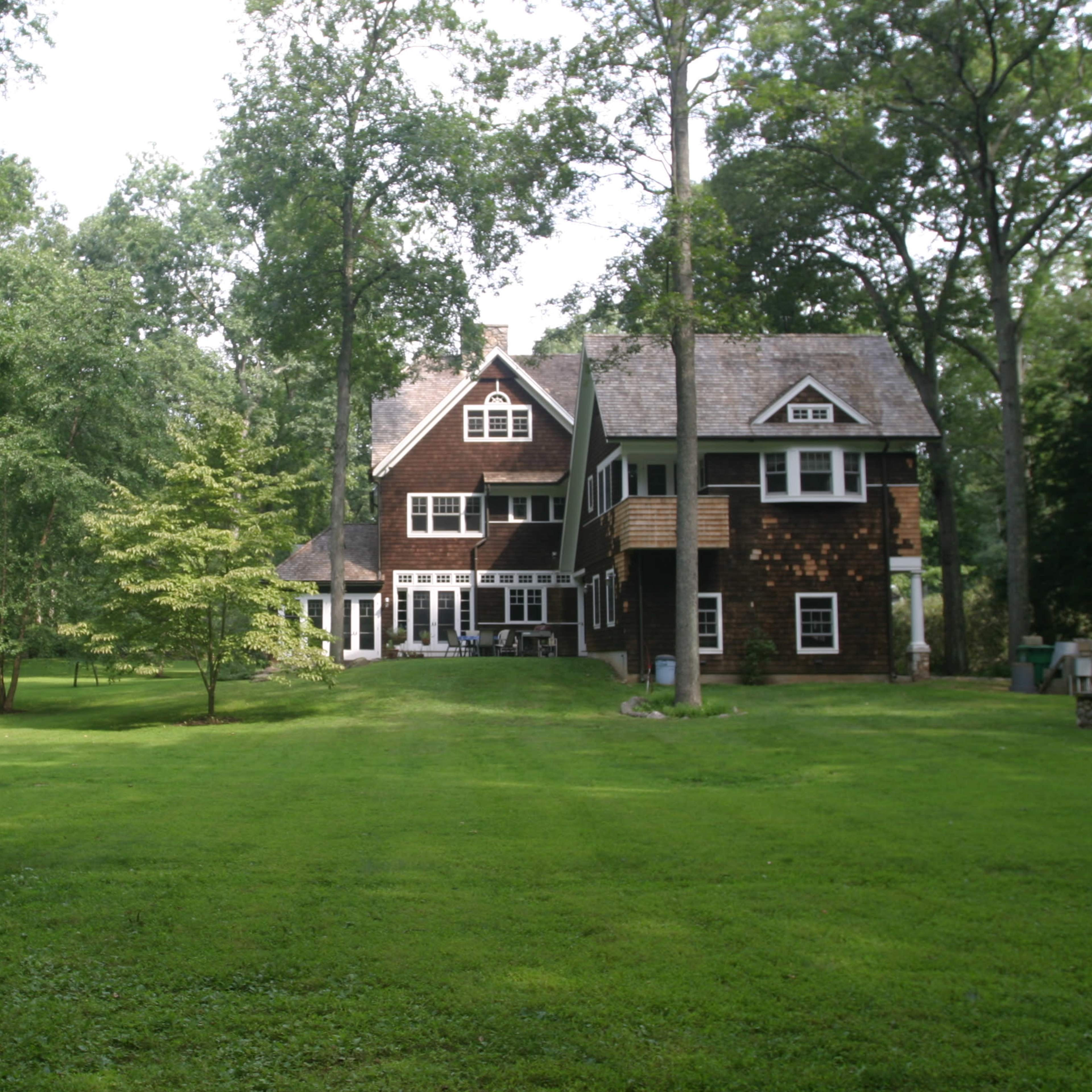 A large, multi-story wooden house is set among tall trees on a well-maintained lawn.