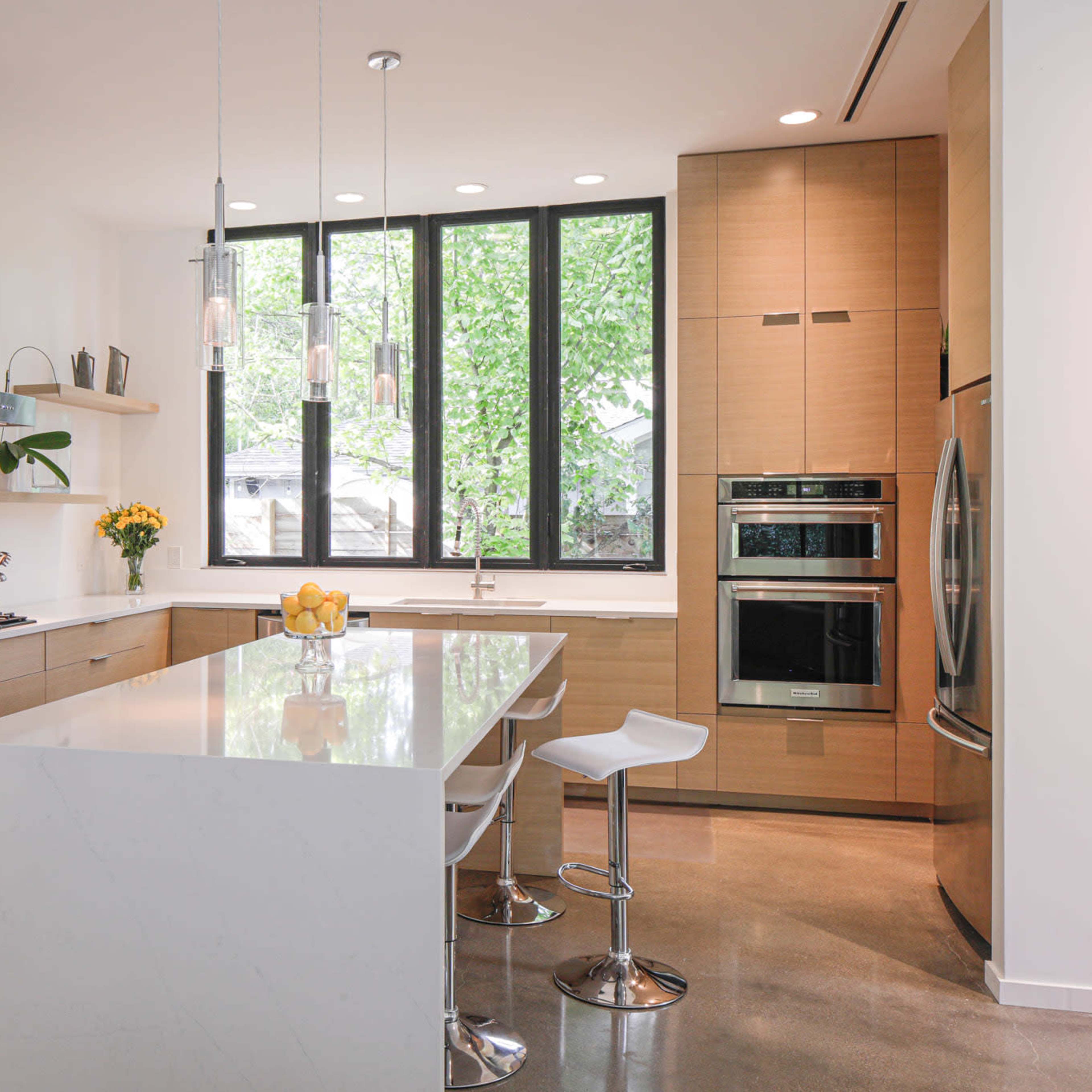 A modern kitchen features a large island with bar stools, sleek cabinetry, and large windows that allow natural light to fill the space.
