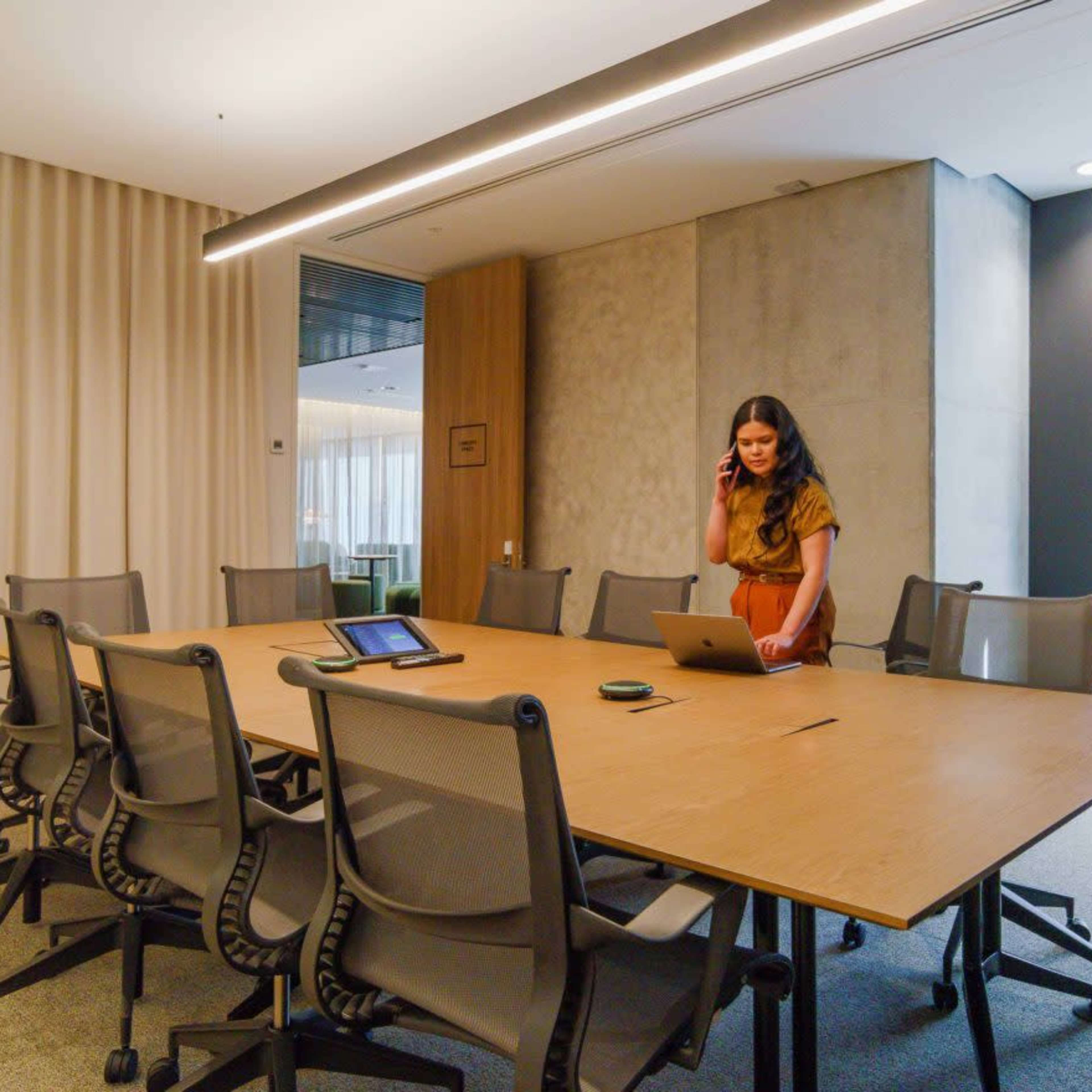 A woman stands in a conference room with a large wooden table surrounded by chairs, working on a laptop while holding a phone to her ear.