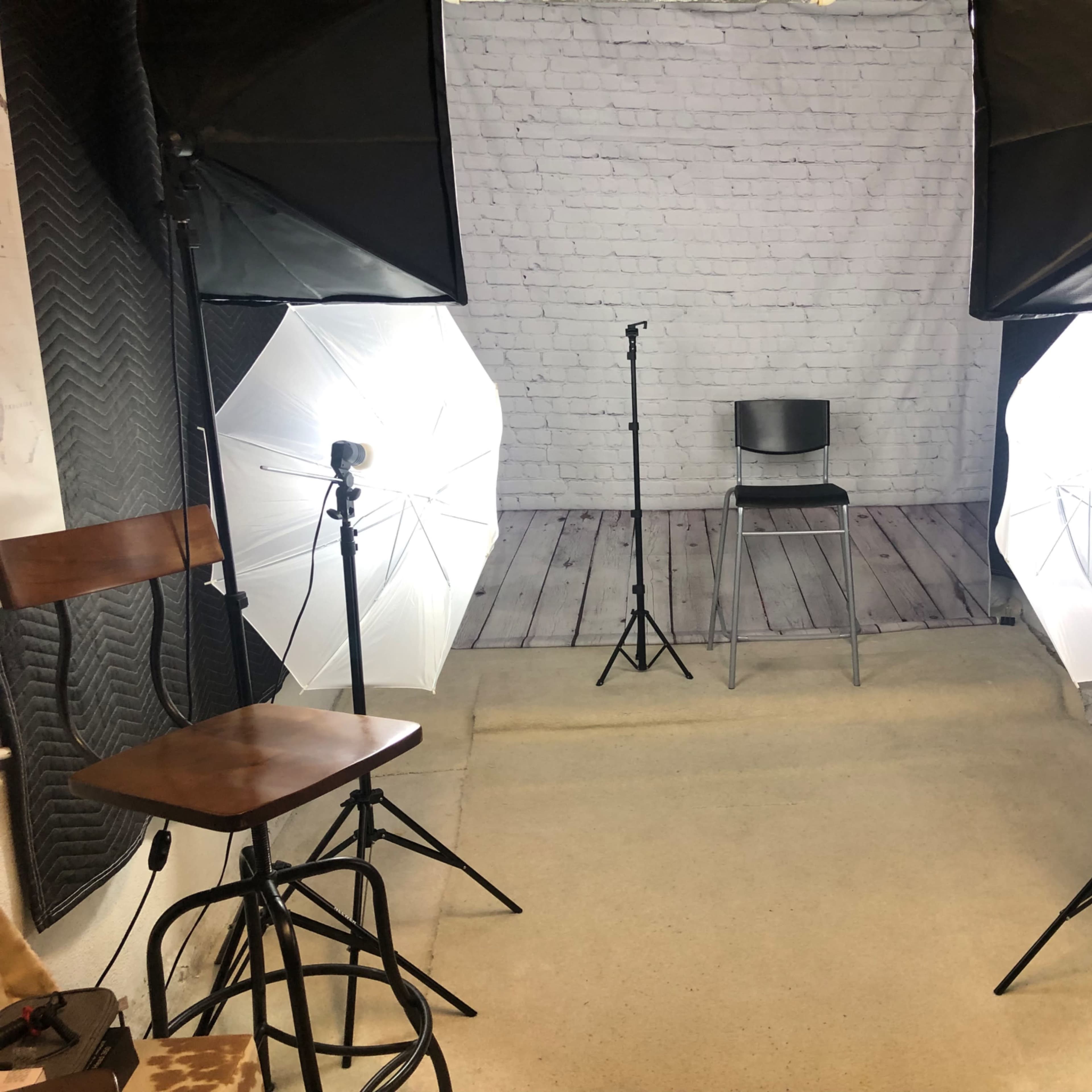 A photo studio setup with two light stands, a black chair in the center, and a backdrop of white brick walls.