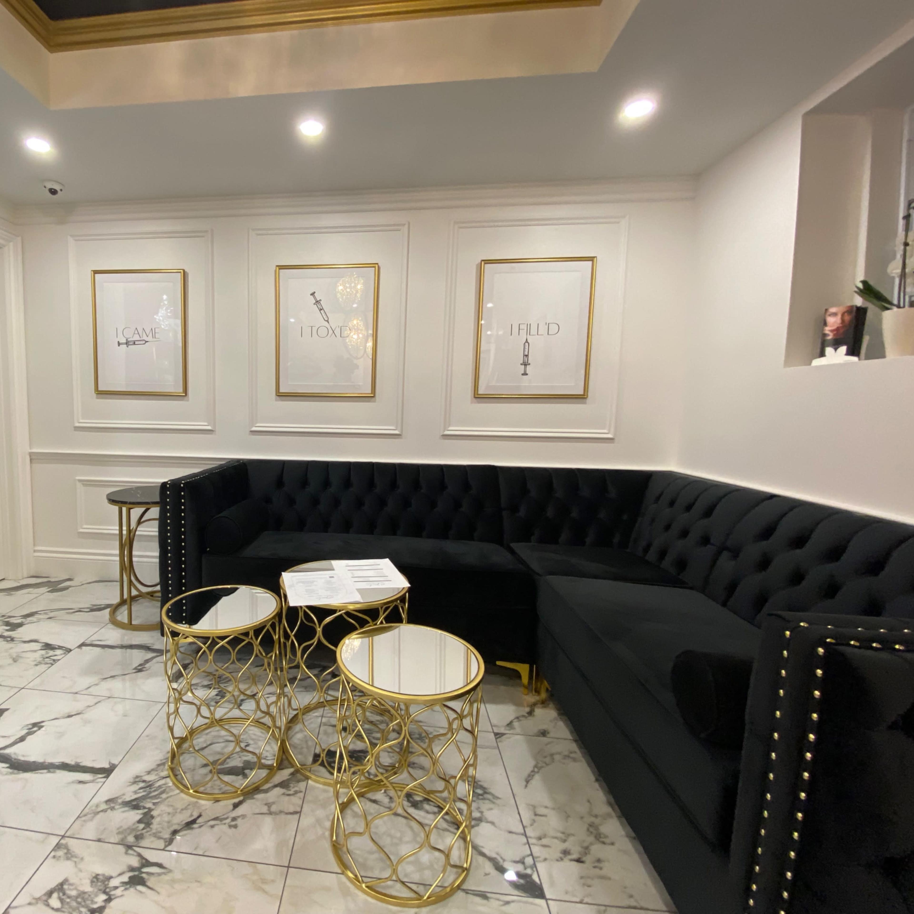 A modern waiting area with a black velvet sofa, marble flooring, and gold-accented tables, along with framed artwork on the walls.