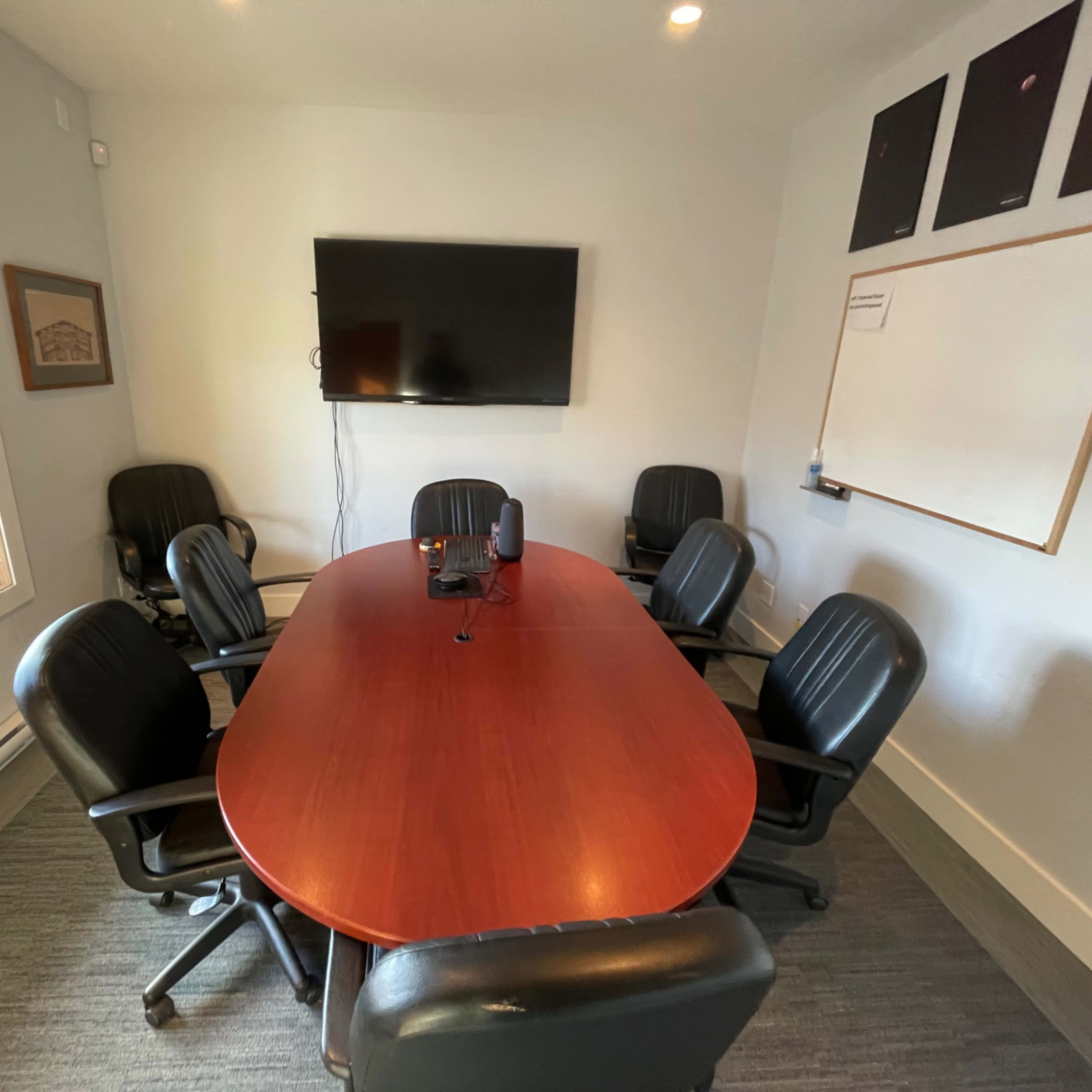 A conference room features a large oval table surrounded by six black chairs, with a wall-mounted television and a whiteboard.