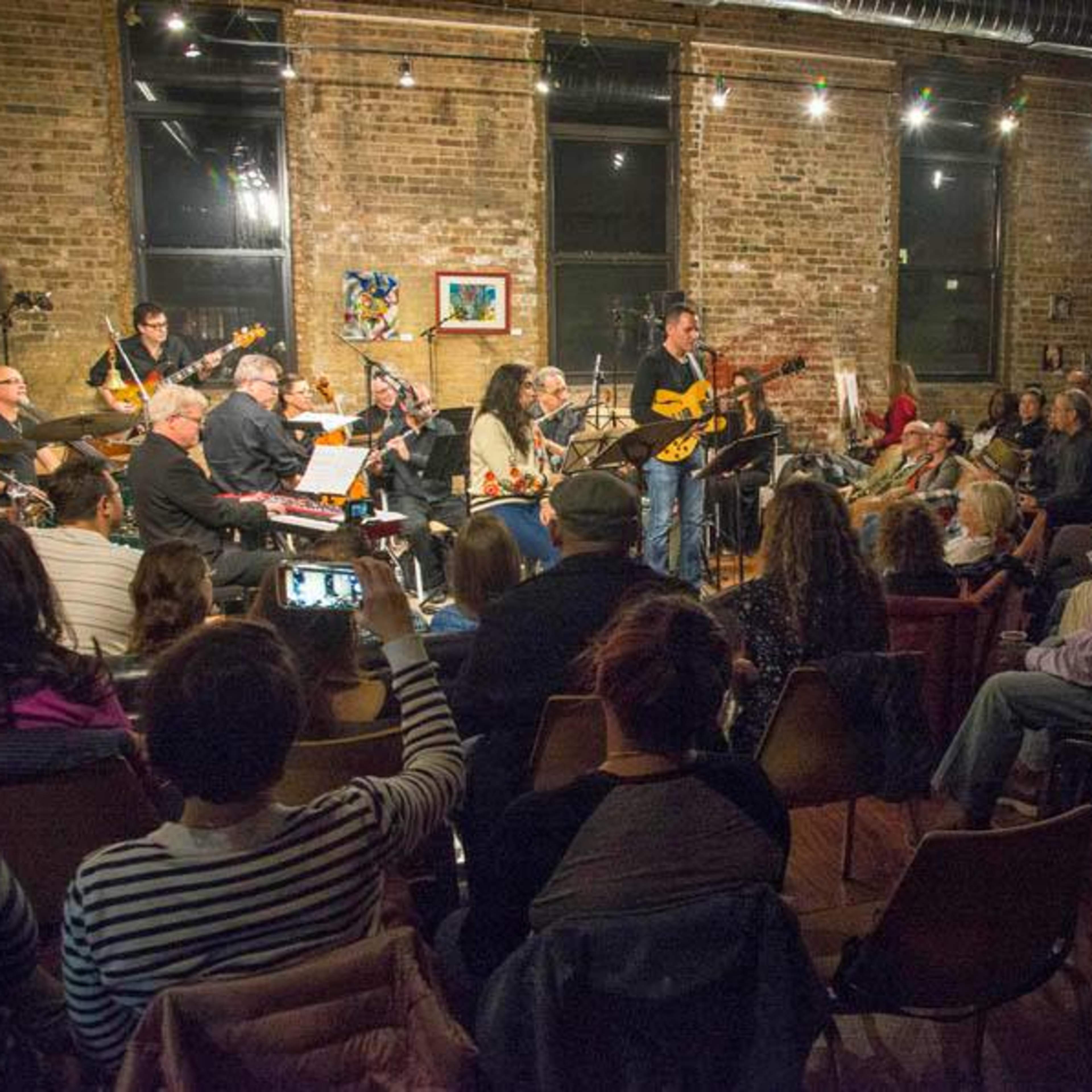 A musician performs on stage in front of a seated audience in a brick-walled venue with various instruments and a band behind him.