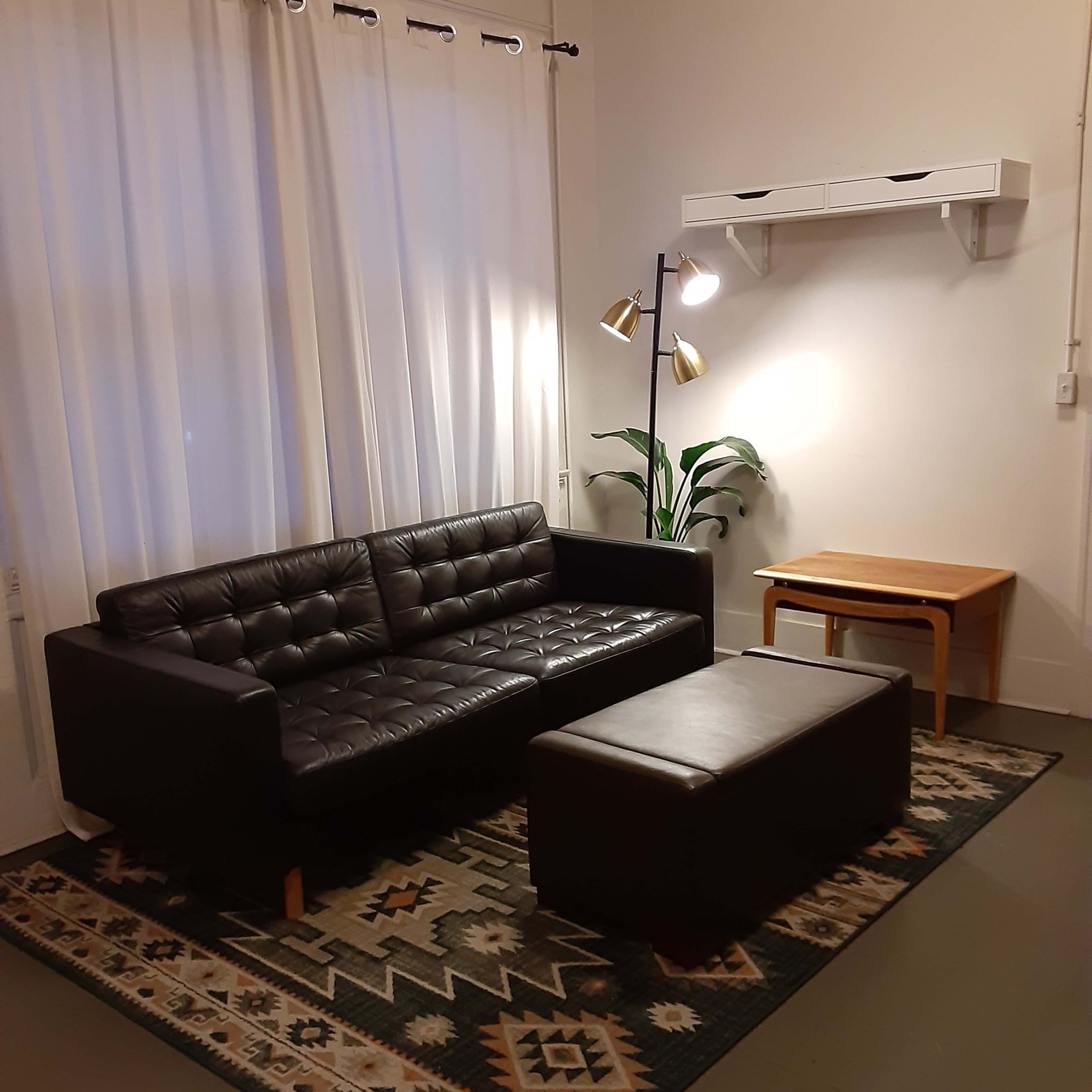 A simple living room featuring a black leather sofa, an ottoman, a small table, and a floor lamp beside a potted plant, all arranged on a patterned rug under large, white curtains.