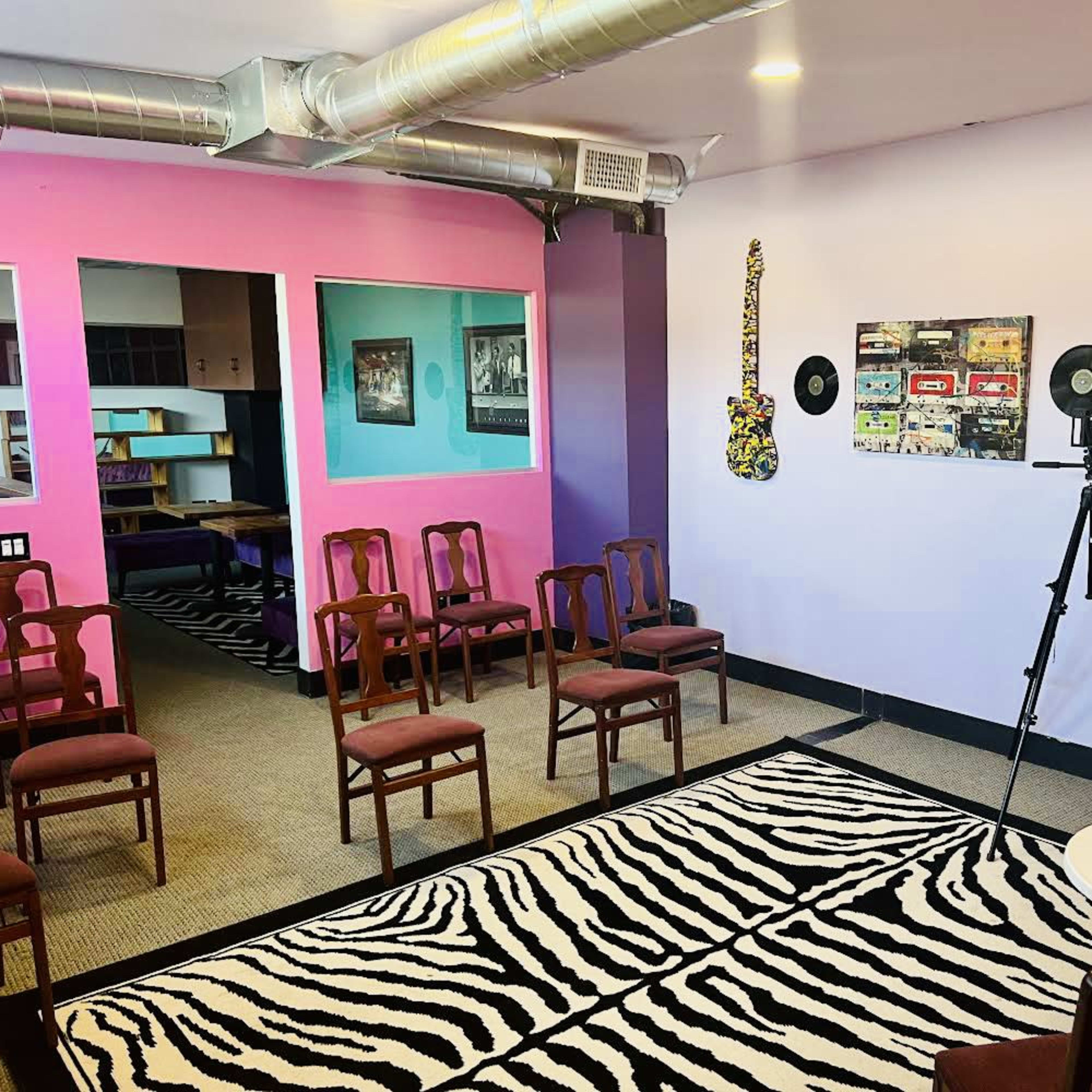 A vibrant room features several chairs arranged in a semi-circle, a camera on a tripod, and decorated walls with colorful artwork and surfboards.