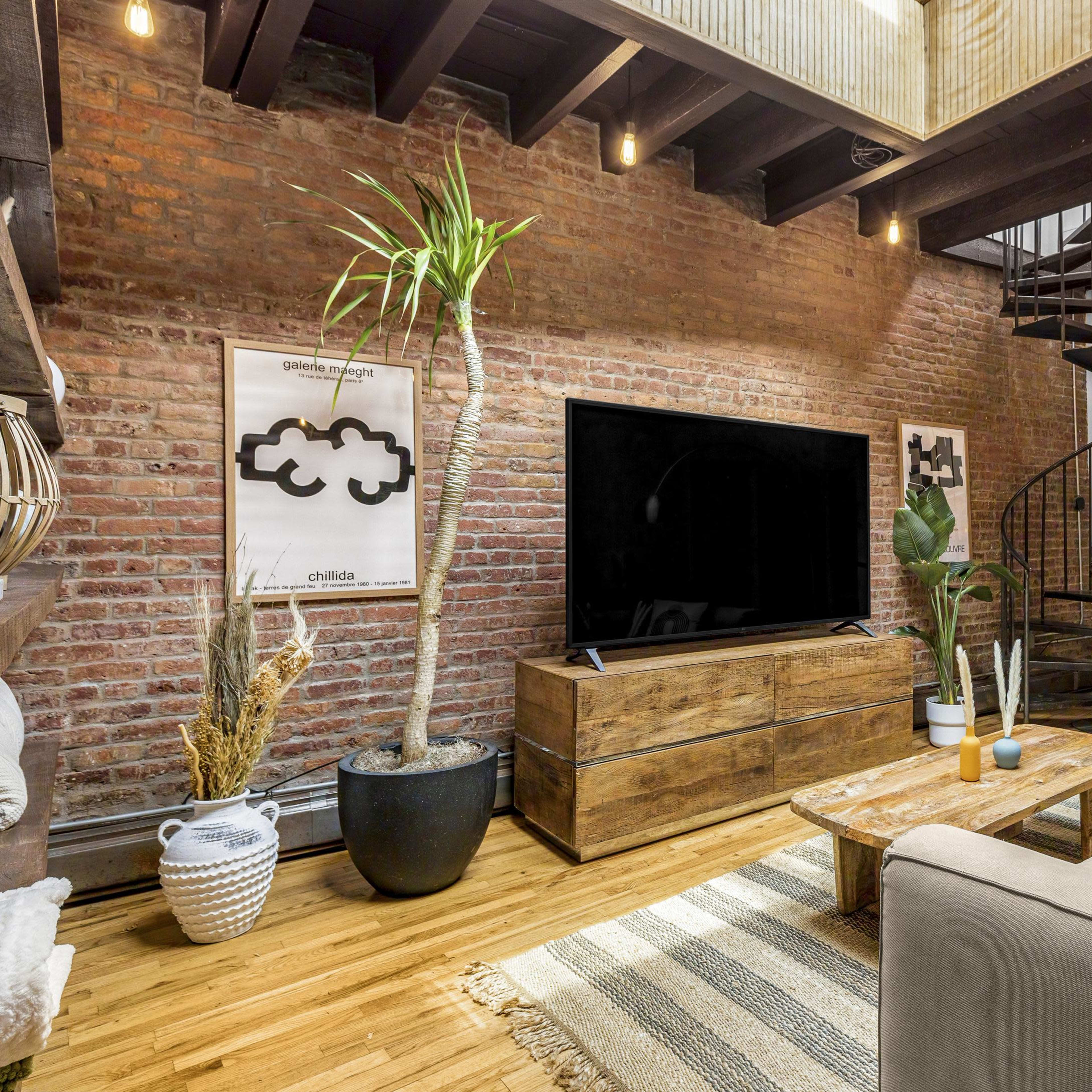 A stylish living room featuring exposed brick walls, a comfortable sofa, a large TV on a wooden stand, and a spiral staircase leading to an upper level.