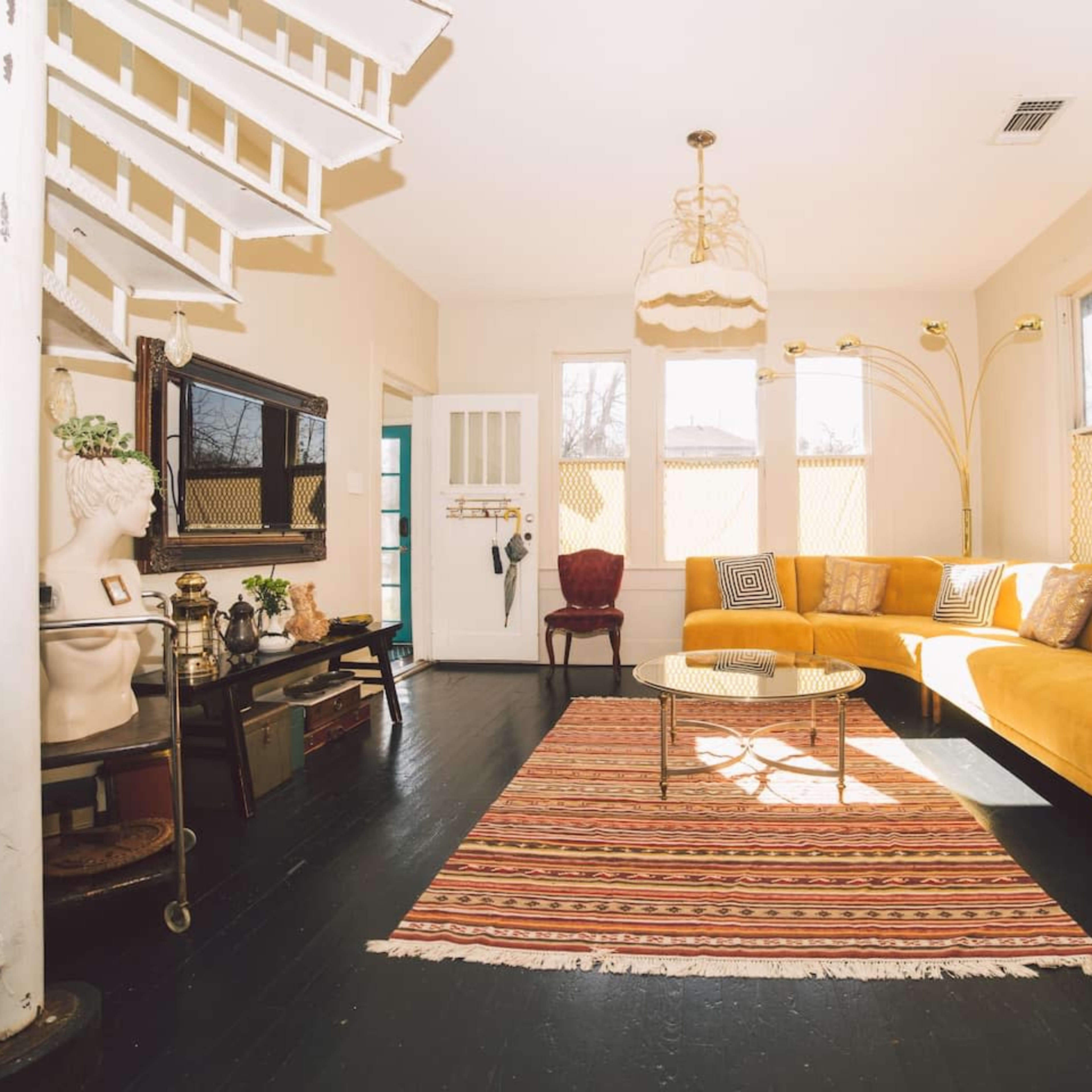 A bright living room features a yellow sectional sofa, a round glass coffee table on a patterned rug, and a staircase with a white railing.