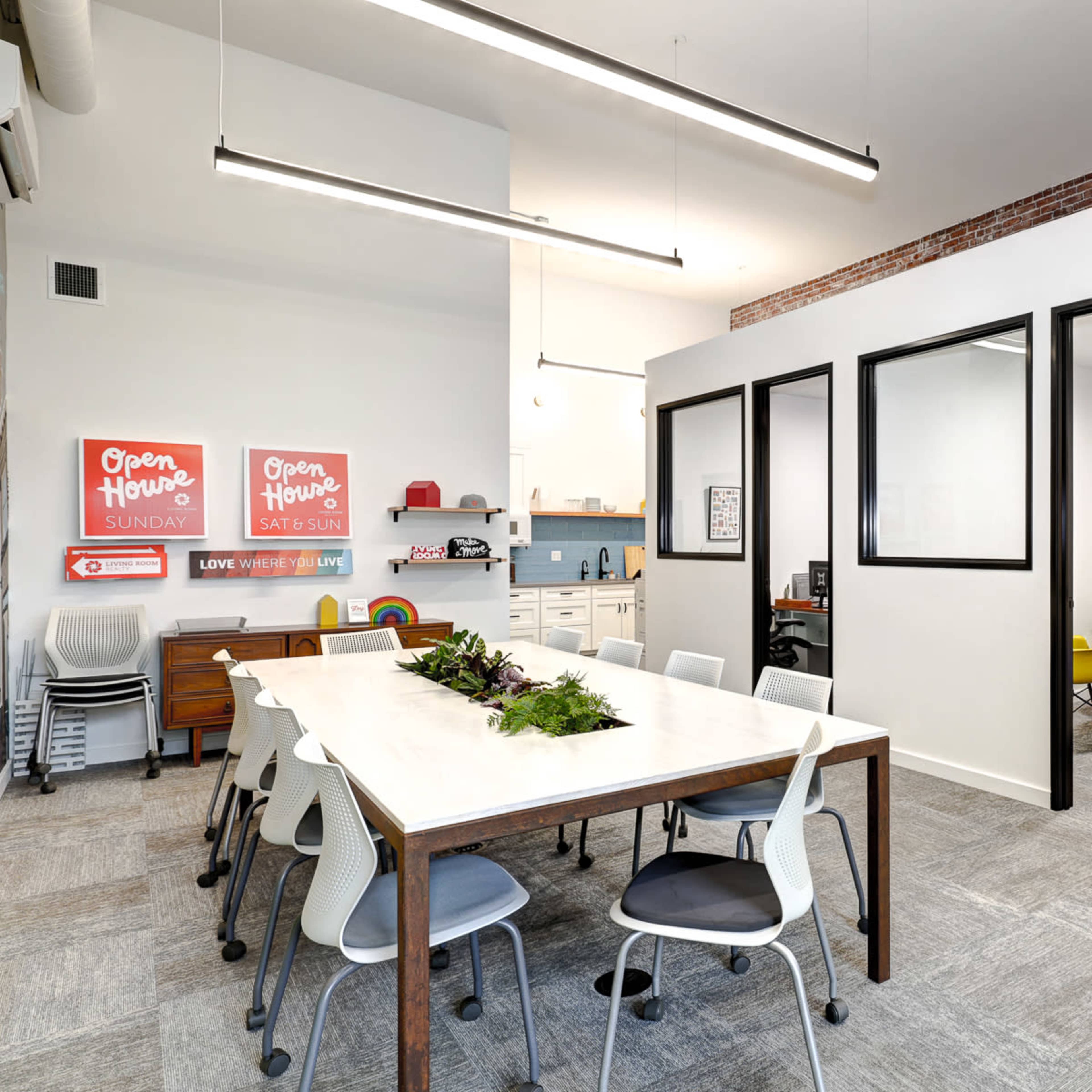 A bright office space features a large table surrounded by chairs, with a kitchenette area and glass-partitioned meeting rooms in the background.