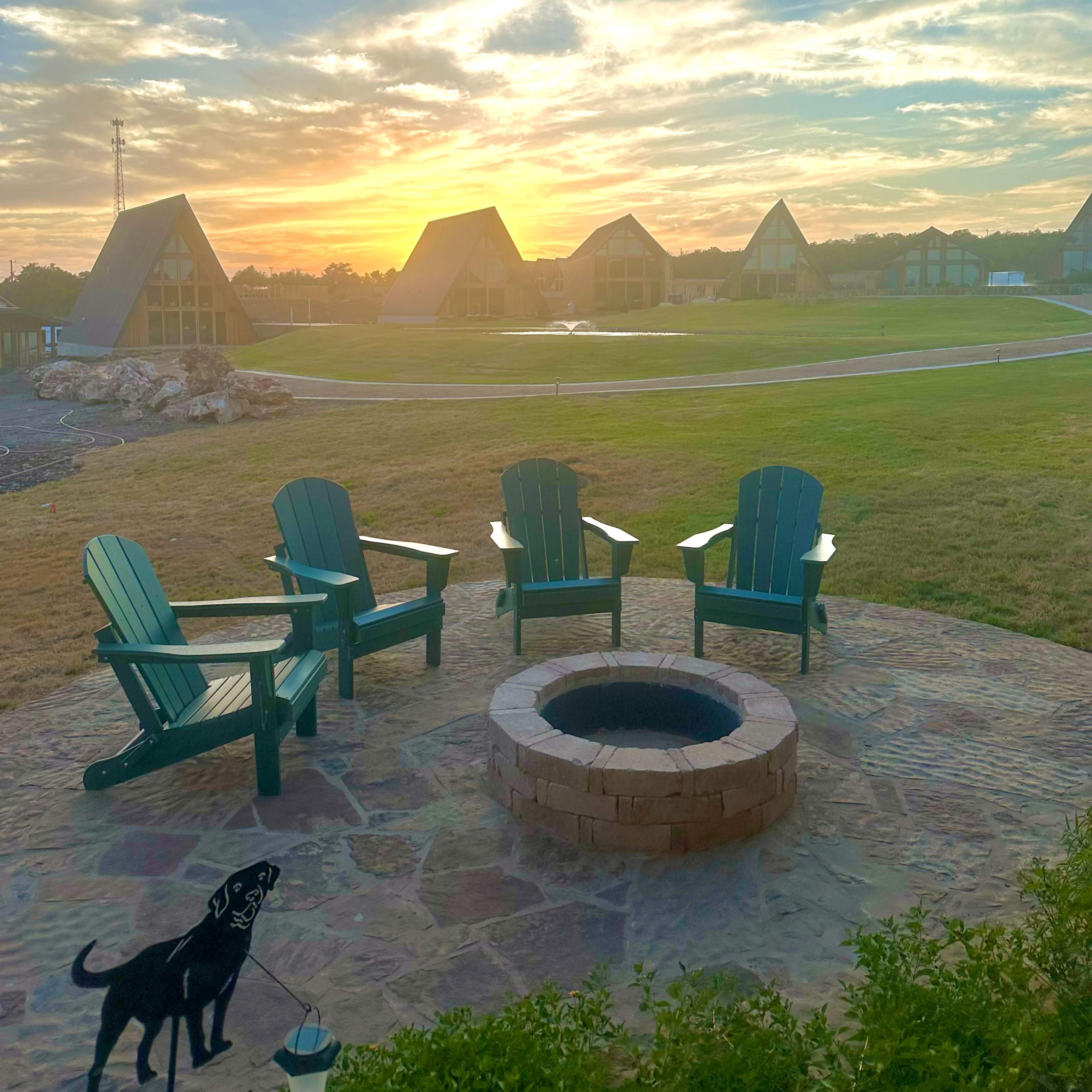 The scene shows a group of four green Adirondack chairs arranged around a circular stone fire pit, with a sunset illuminating the sky over several triangular-roofed buildings in the background.