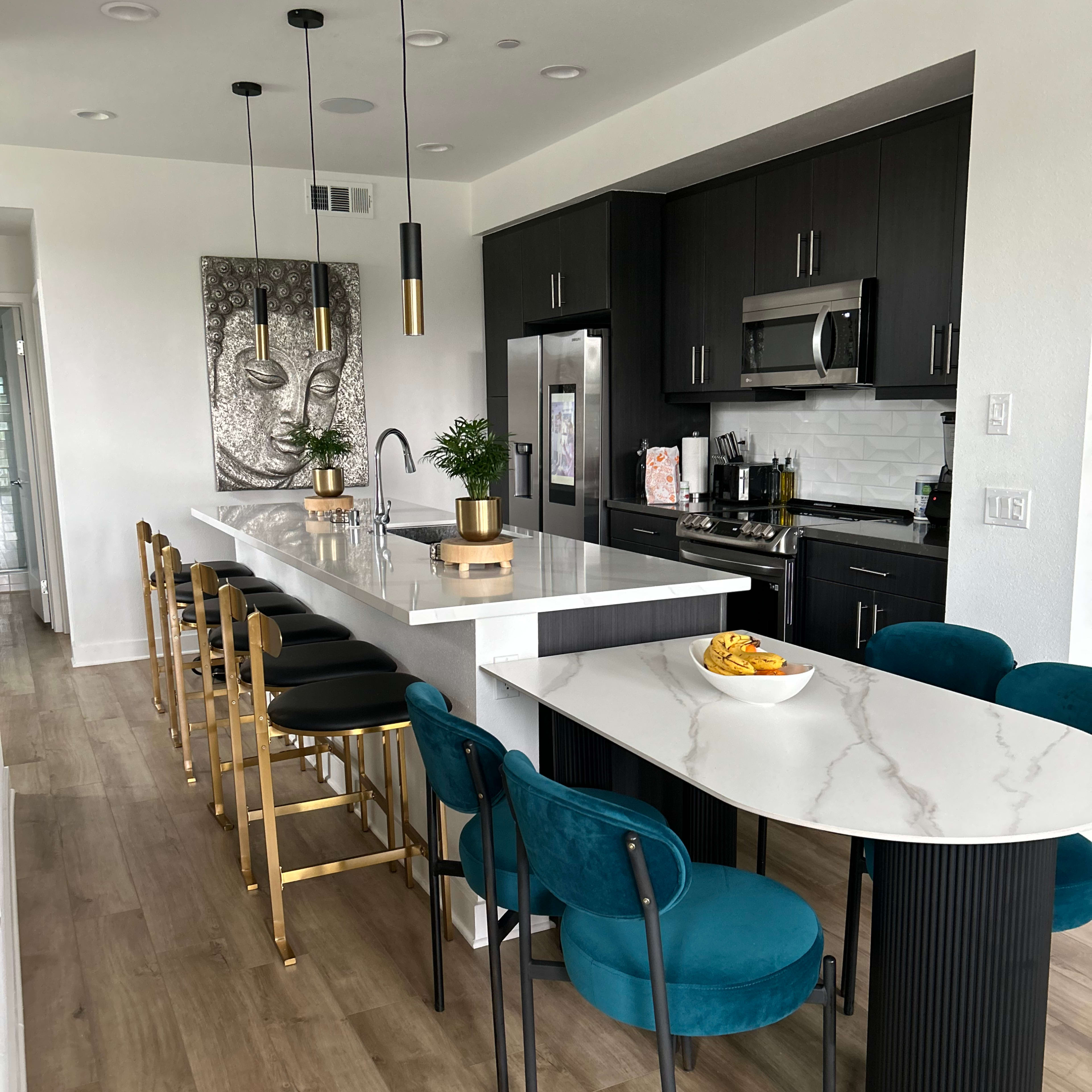 A modern kitchen with dark cabinets, a large island with white marble countertops, and stylish bar stools, along with a round dining table and teal upholstered chairs.