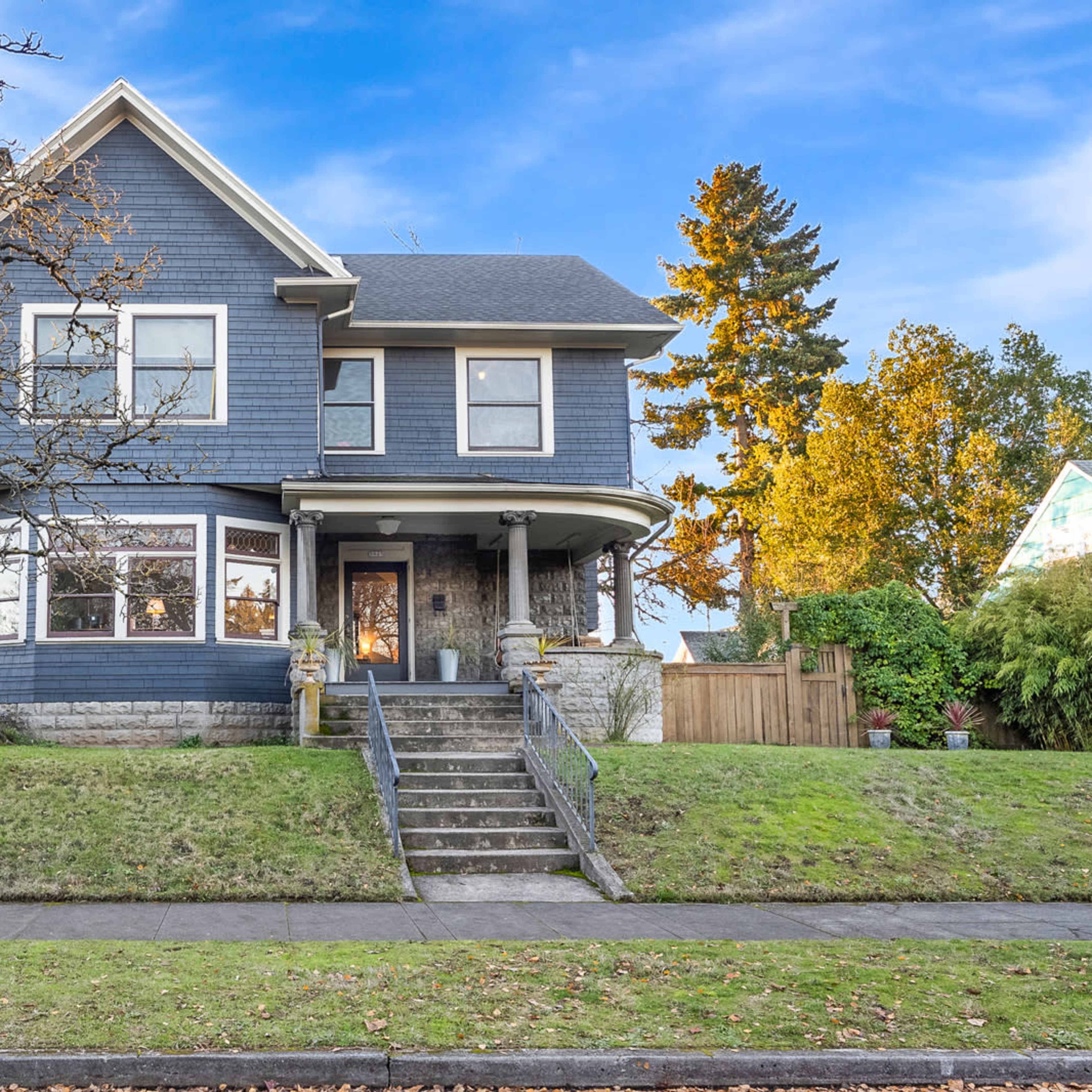 A two-story blue house with a front porch and large windows is situated on a green lawn, bordered by a tree and a sidewalk.