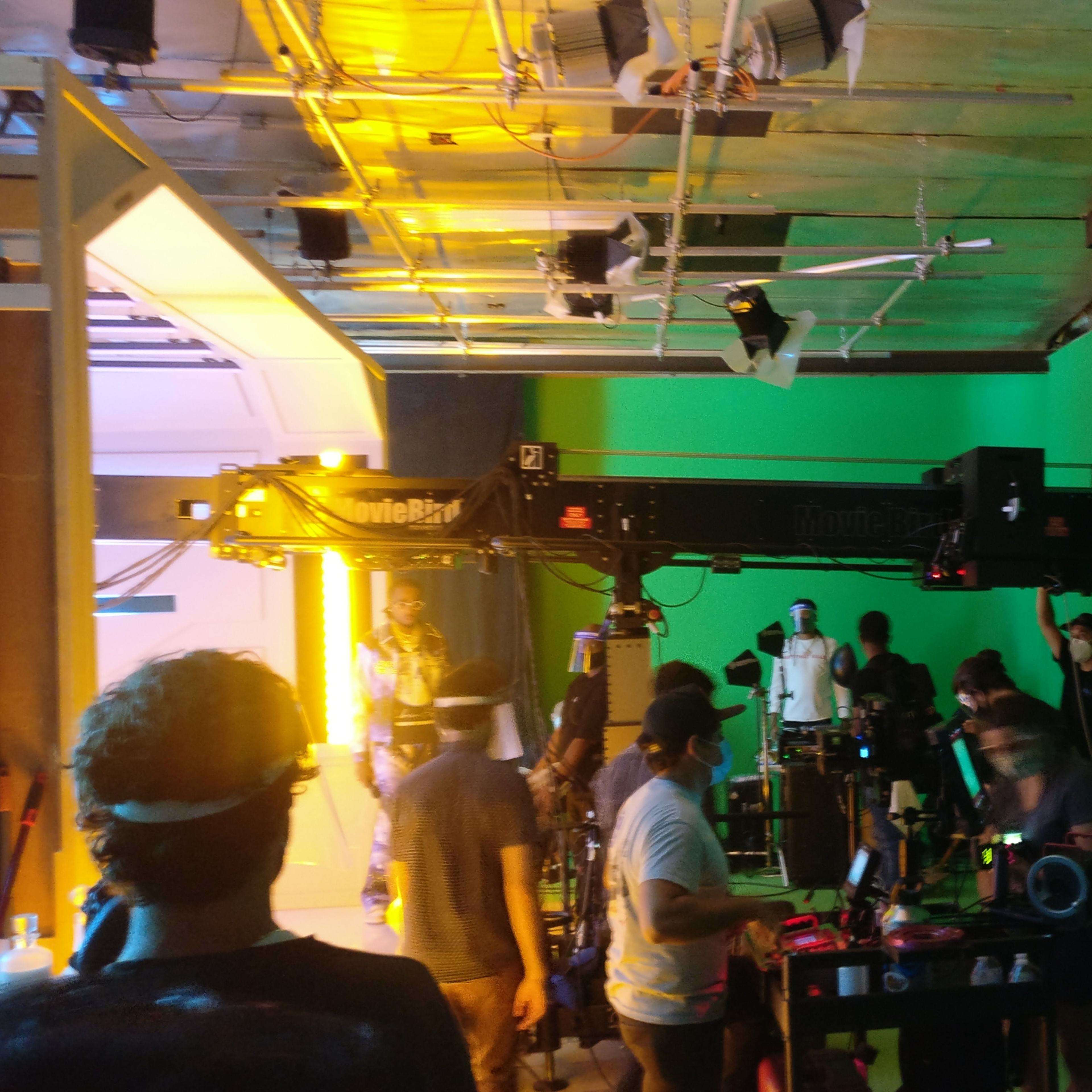 A film crew operates various equipment in a studio with green walls and bright lighting.