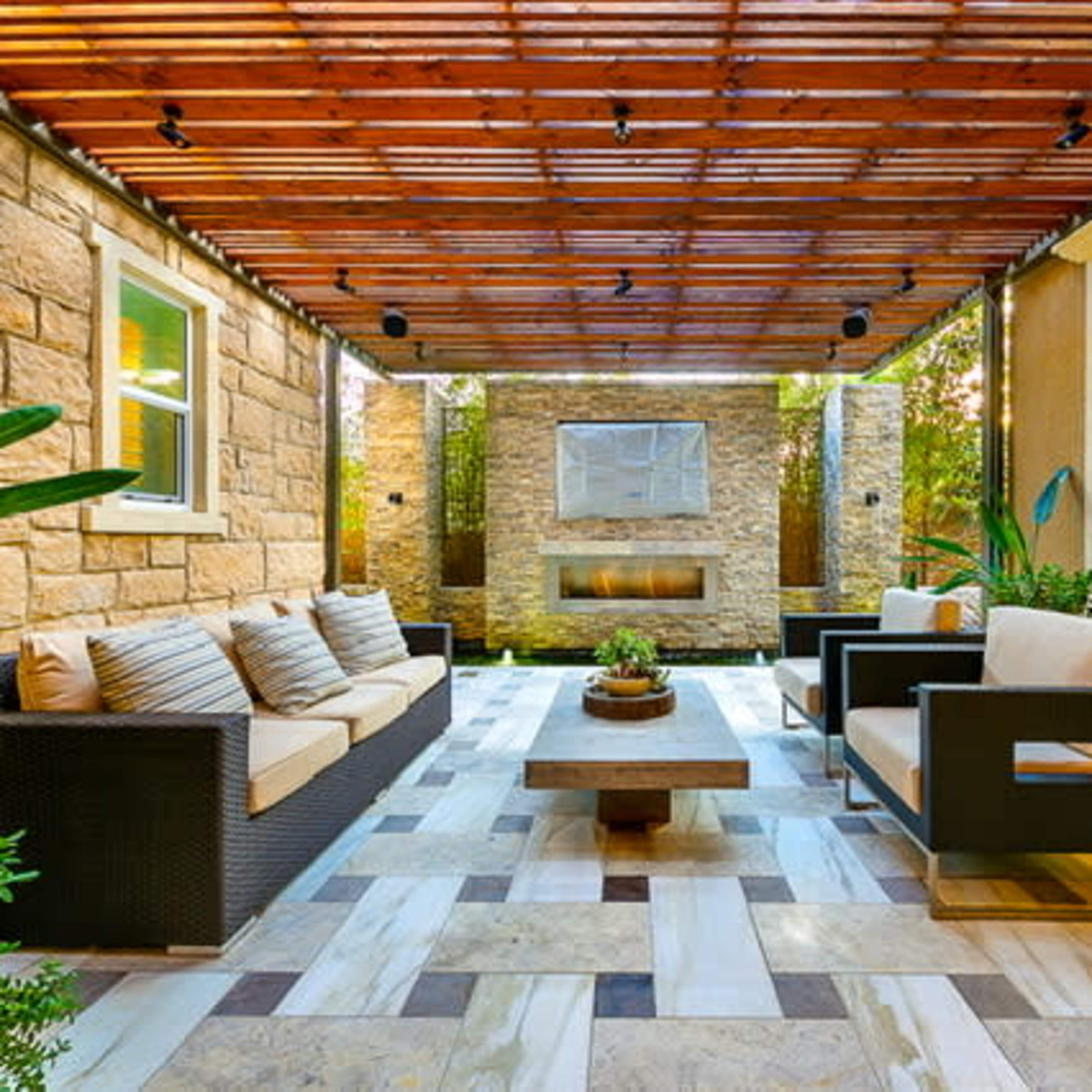 A covered patio features a seating area with a couch, two chairs, and a coffee table, surrounded by stone walls and greenery.