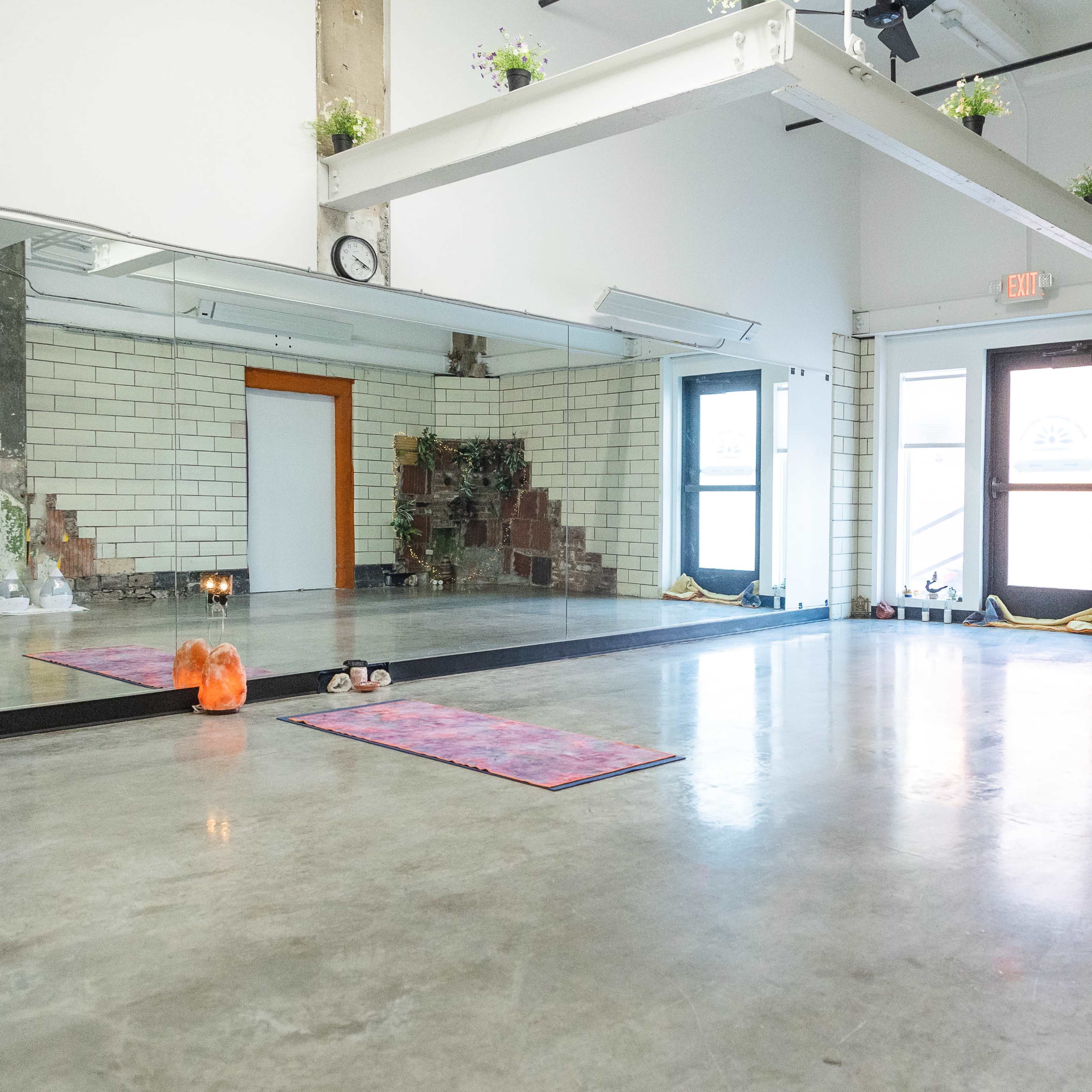 A spacious, empty yoga studio with a mirror and yoga mat on the floor, featuring brick walls and large windows.