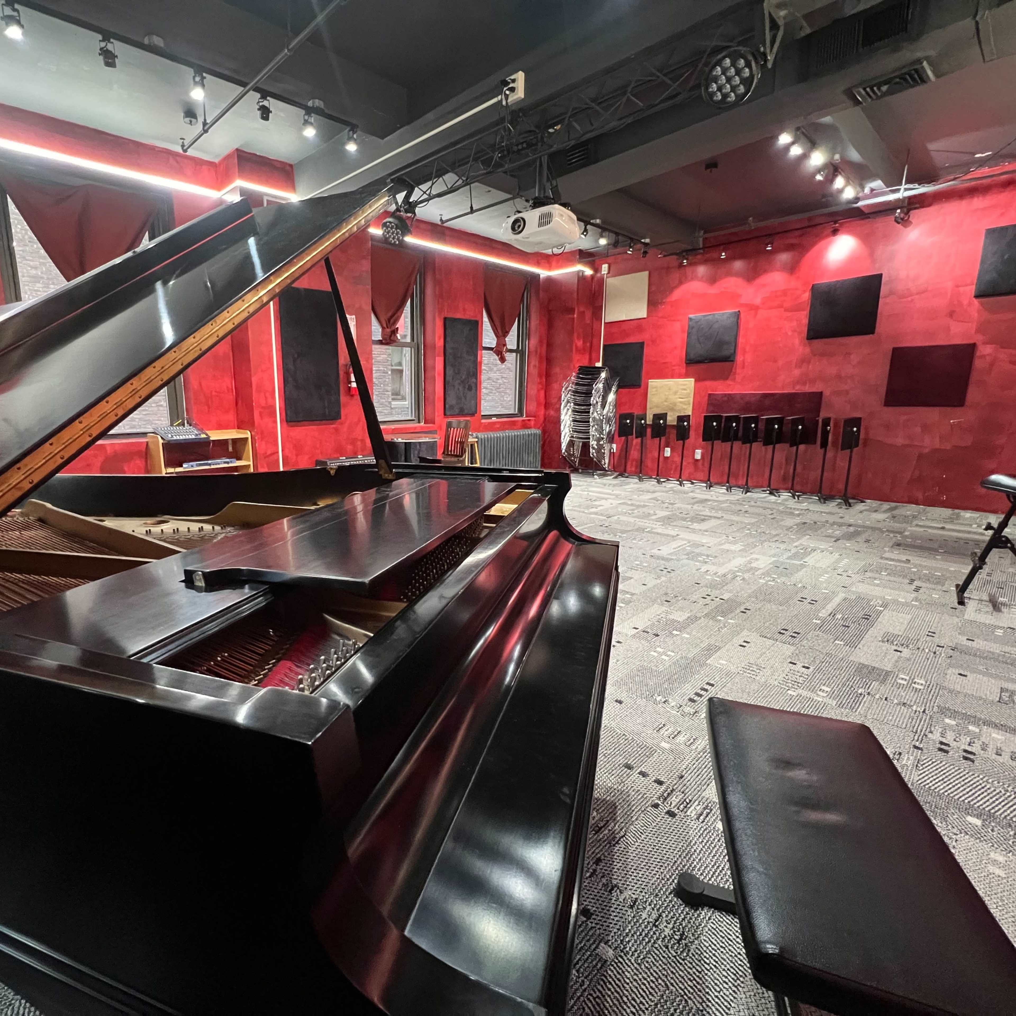 A grand piano in a music studio with red walls and soundproof panels, equipped with audio equipment.