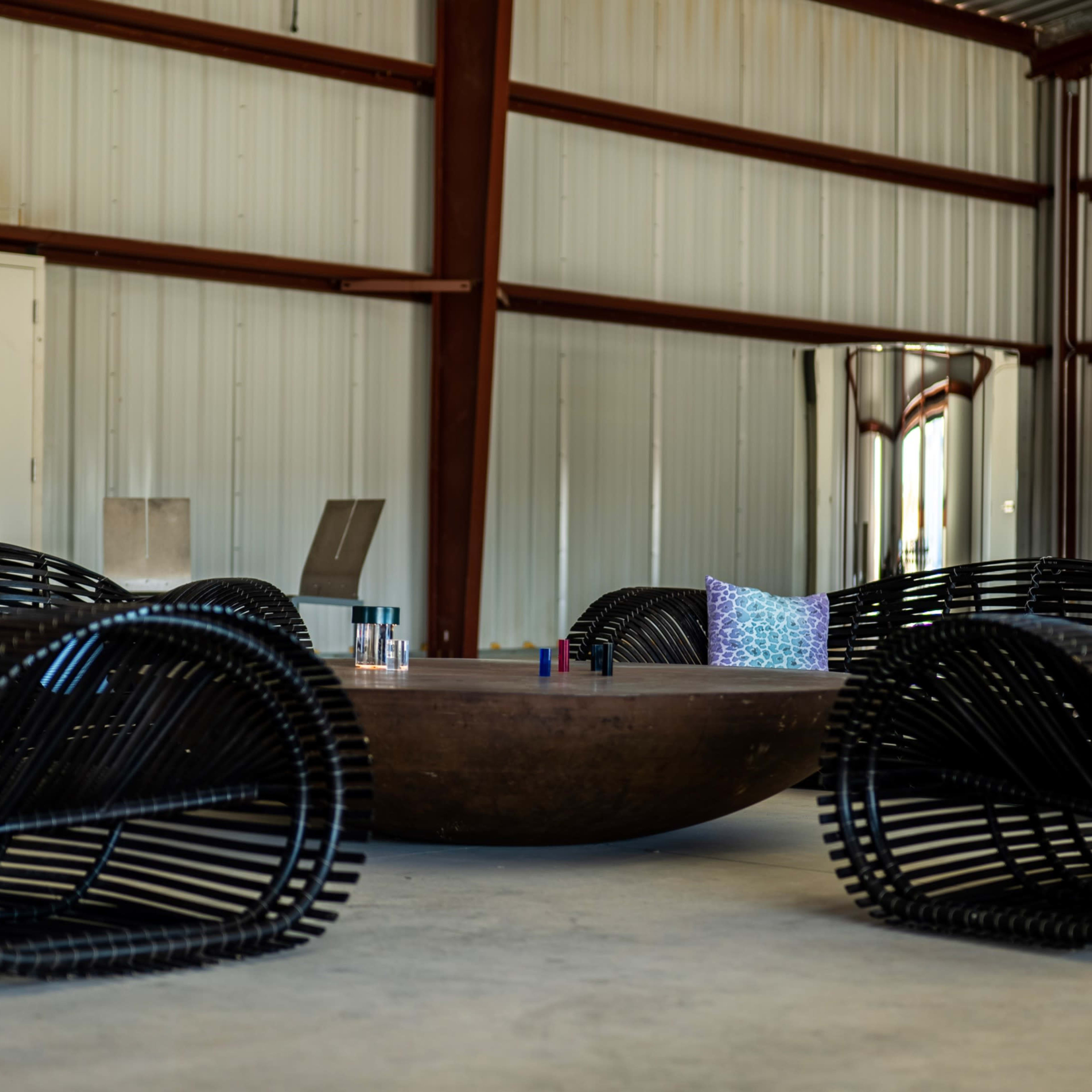 A circular wooden table is surrounded by four black wicker chairs within a metal-clad building.