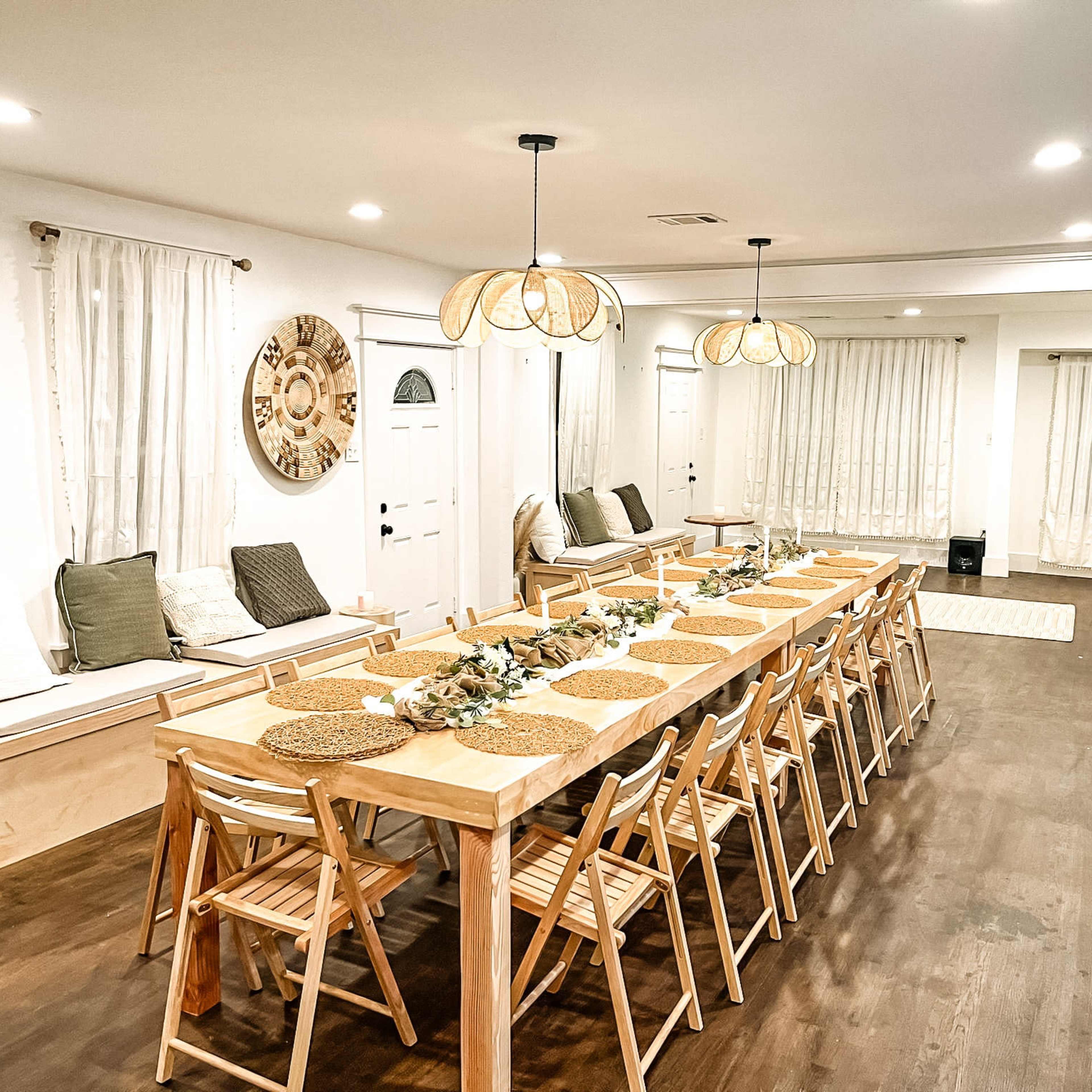 A long wooden dining table with woven placemats is set for a meal in a well-lit dining room featuring white walls, large windows with sheer curtains, and a cozy seating area.
