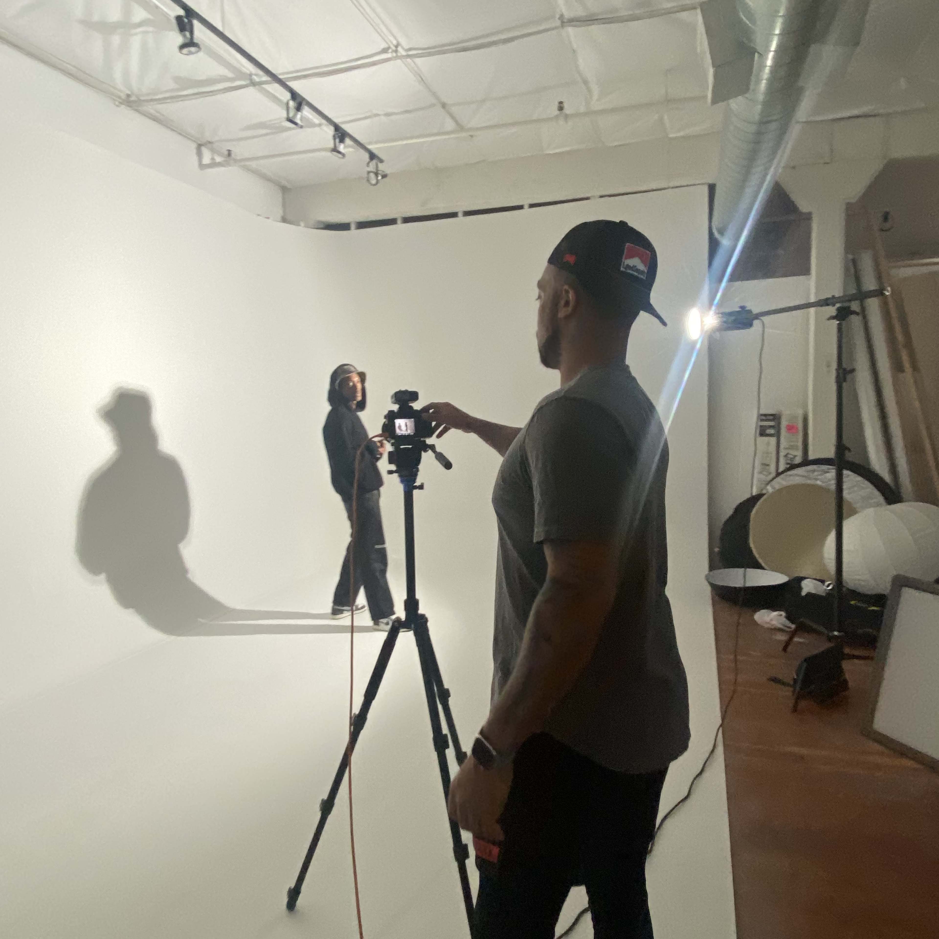 A person adjusts a camera on a tripod while another person poses in front of a white backdrop in a photography studio.
