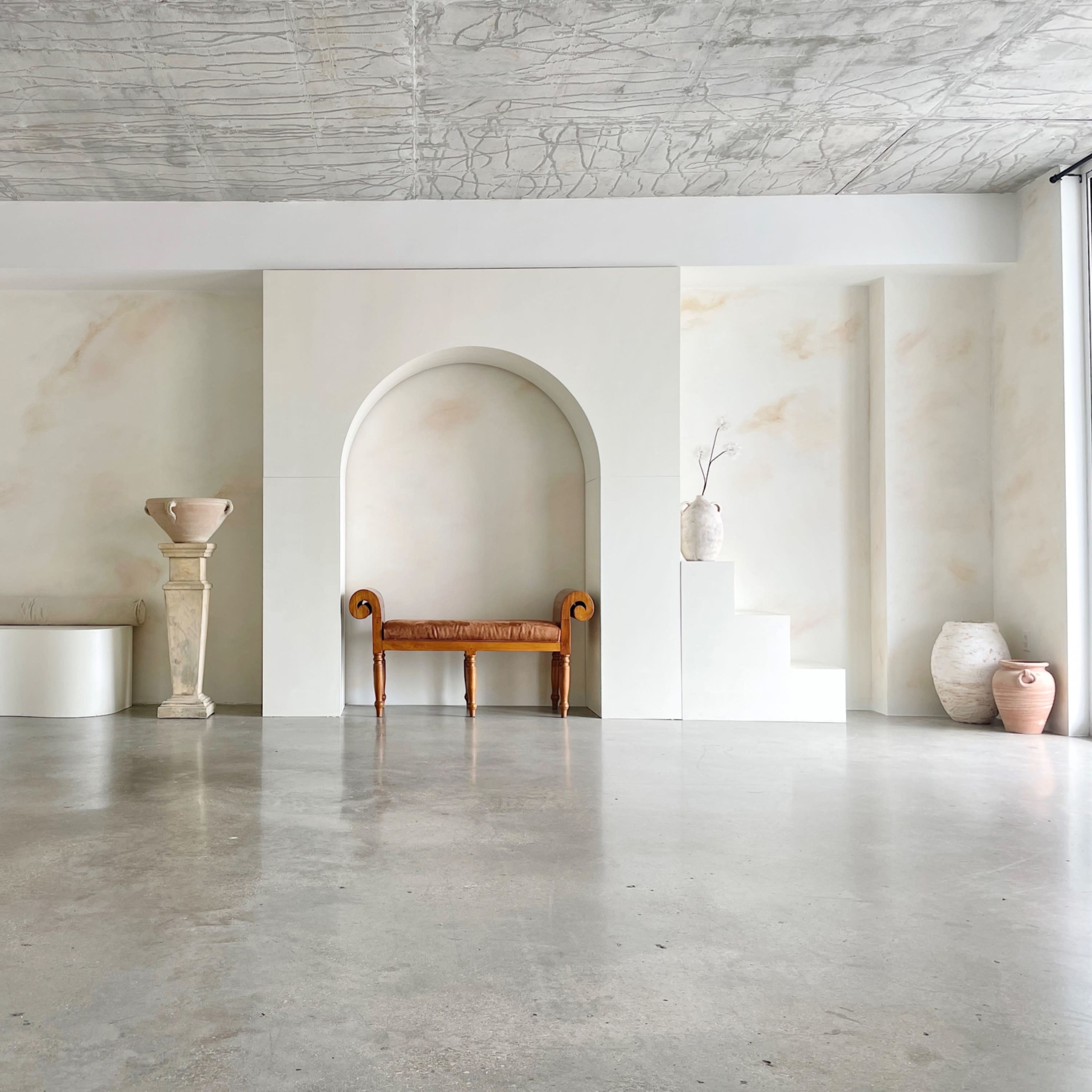 A minimalist interior space featuring a white wall nook with a sculptural vase, a wooden bench, a pedestal with a decorative urn, and a staircase leading to a raised area.