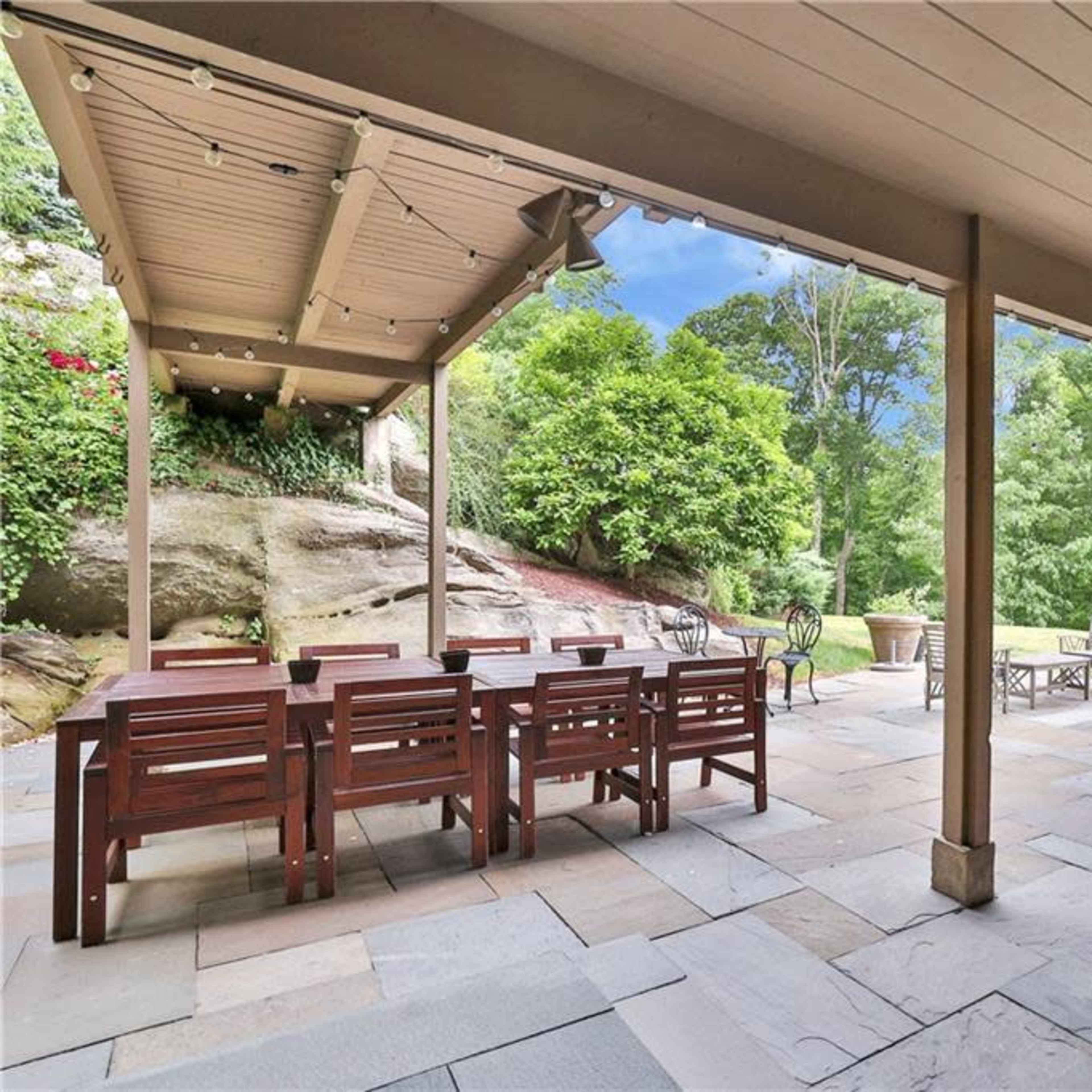 A covered outdoor patio with wooden dining tables and stone flooring, surrounded by greenery and rock formations.
