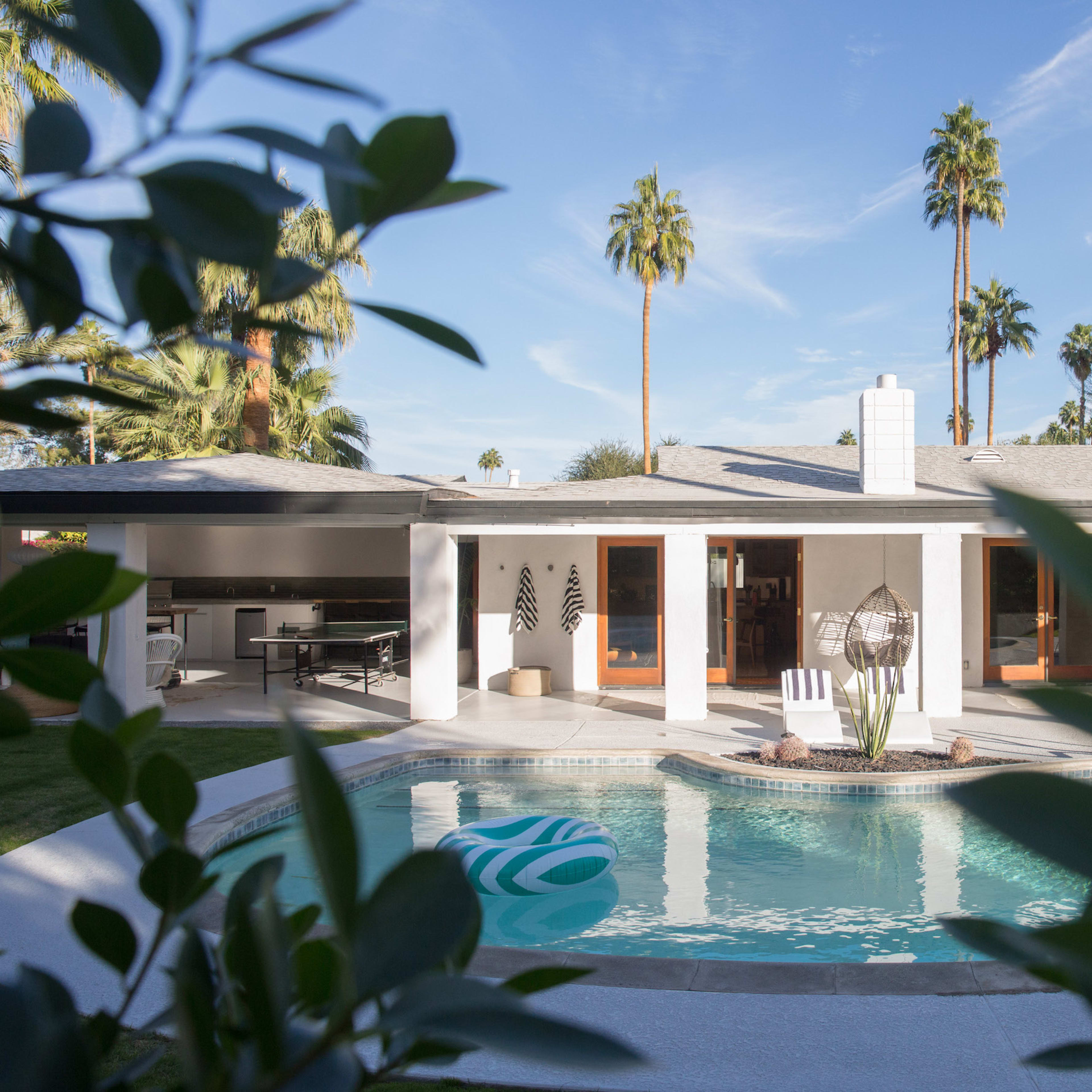 A modern house with large windows overlooks a pool surrounded by palm trees.