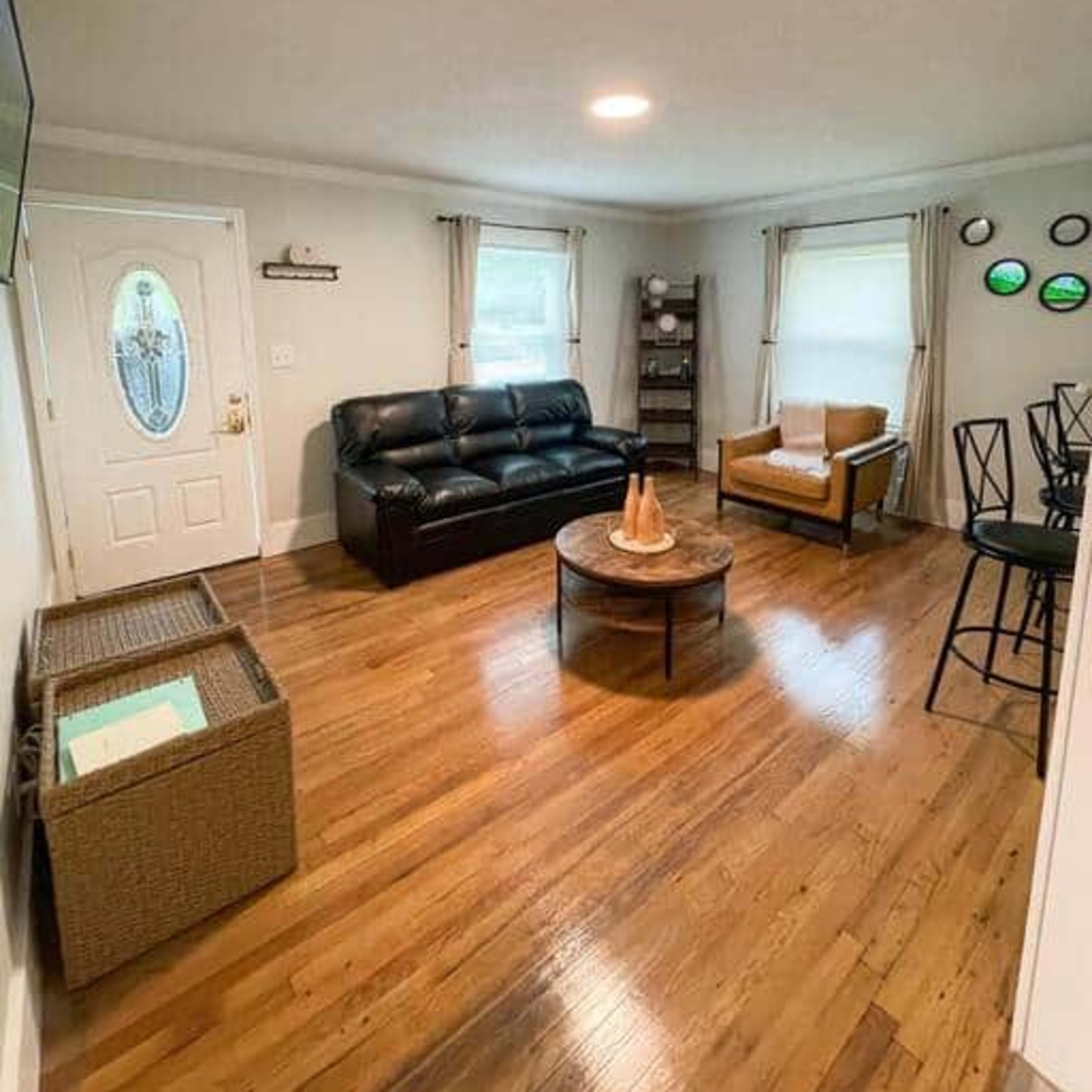 A living room features a black sofa, a brown armchair, a circular wooden coffee table, and a door leading outside.