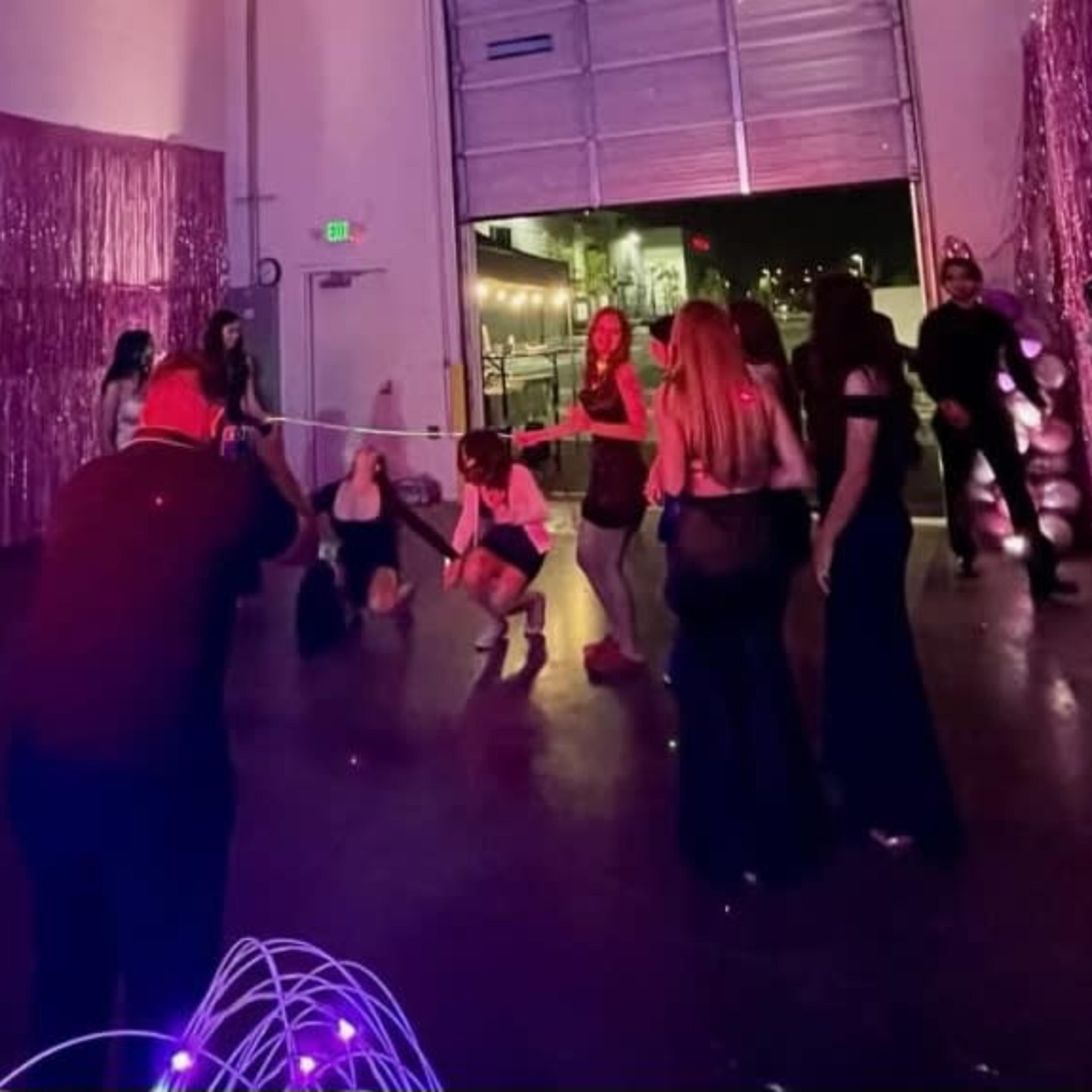 A group of people is participating in a limbo game at a party, with a shiny, pink backdrop and various costumes present.