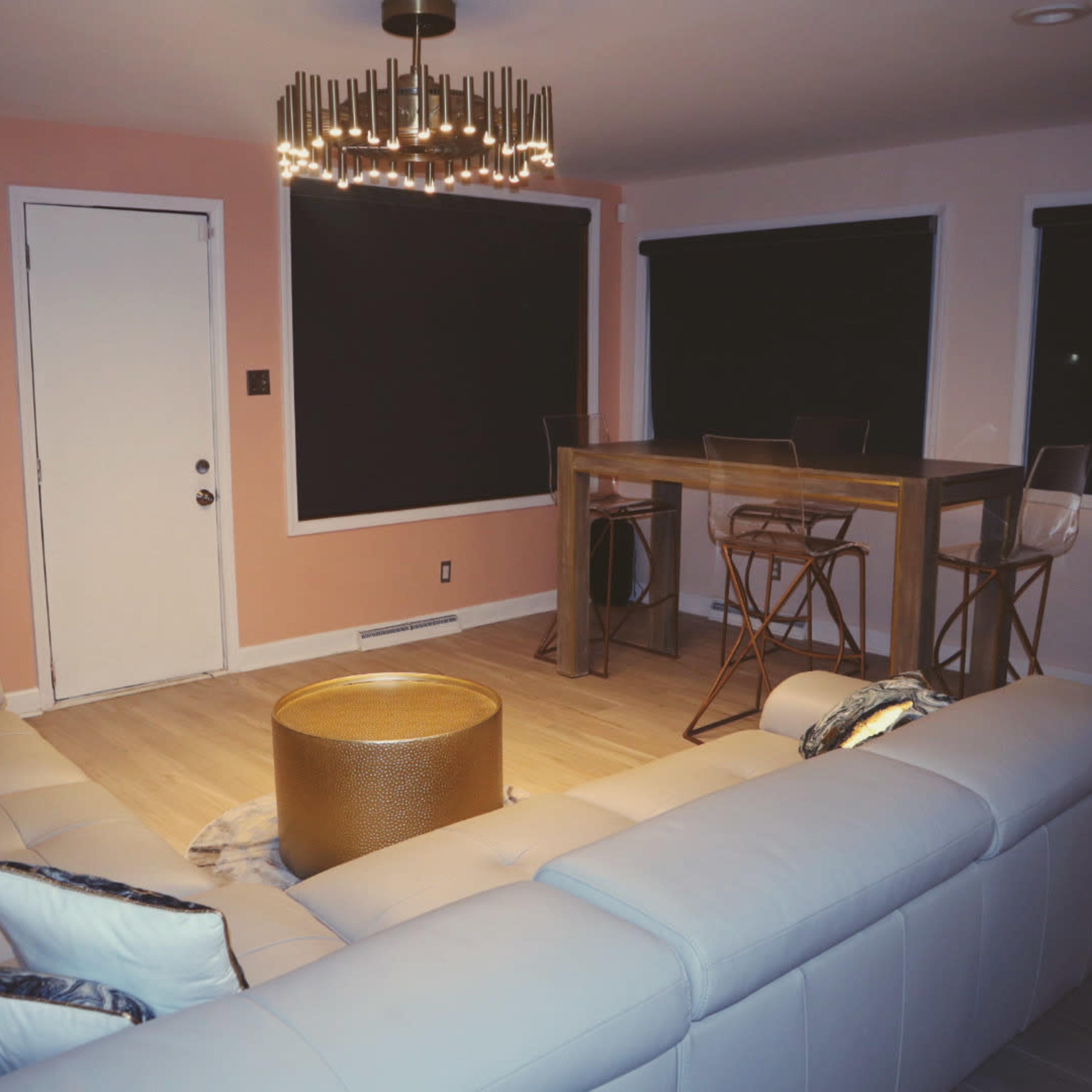The room features a light-colored sectional sofa, a round gold coffee table, and a dining area with a tall table and clear stools, all set against pink and dark walls.