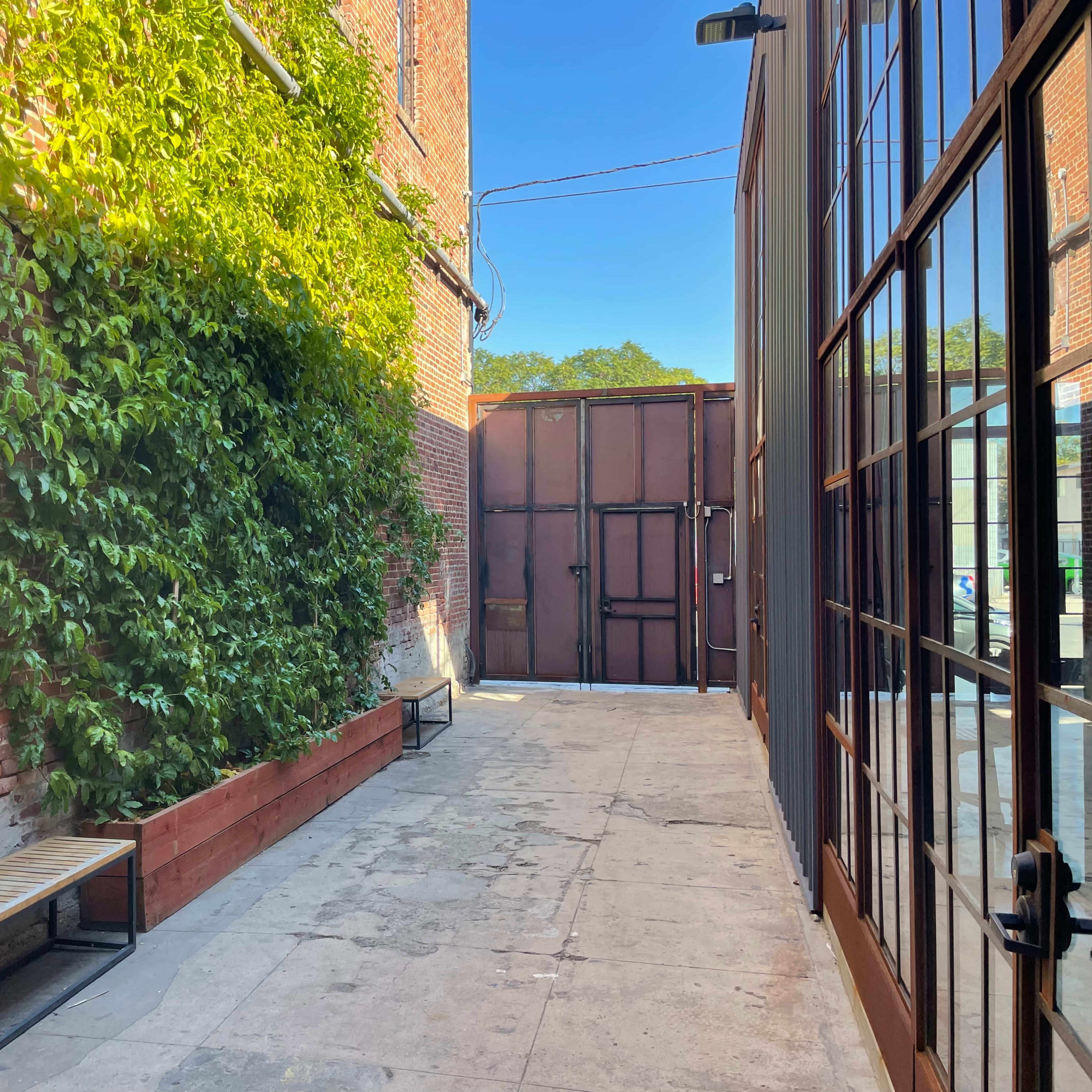 A narrow alleyway features a brick wall with greenery, benches on either side, and a large metal gate at the far end.