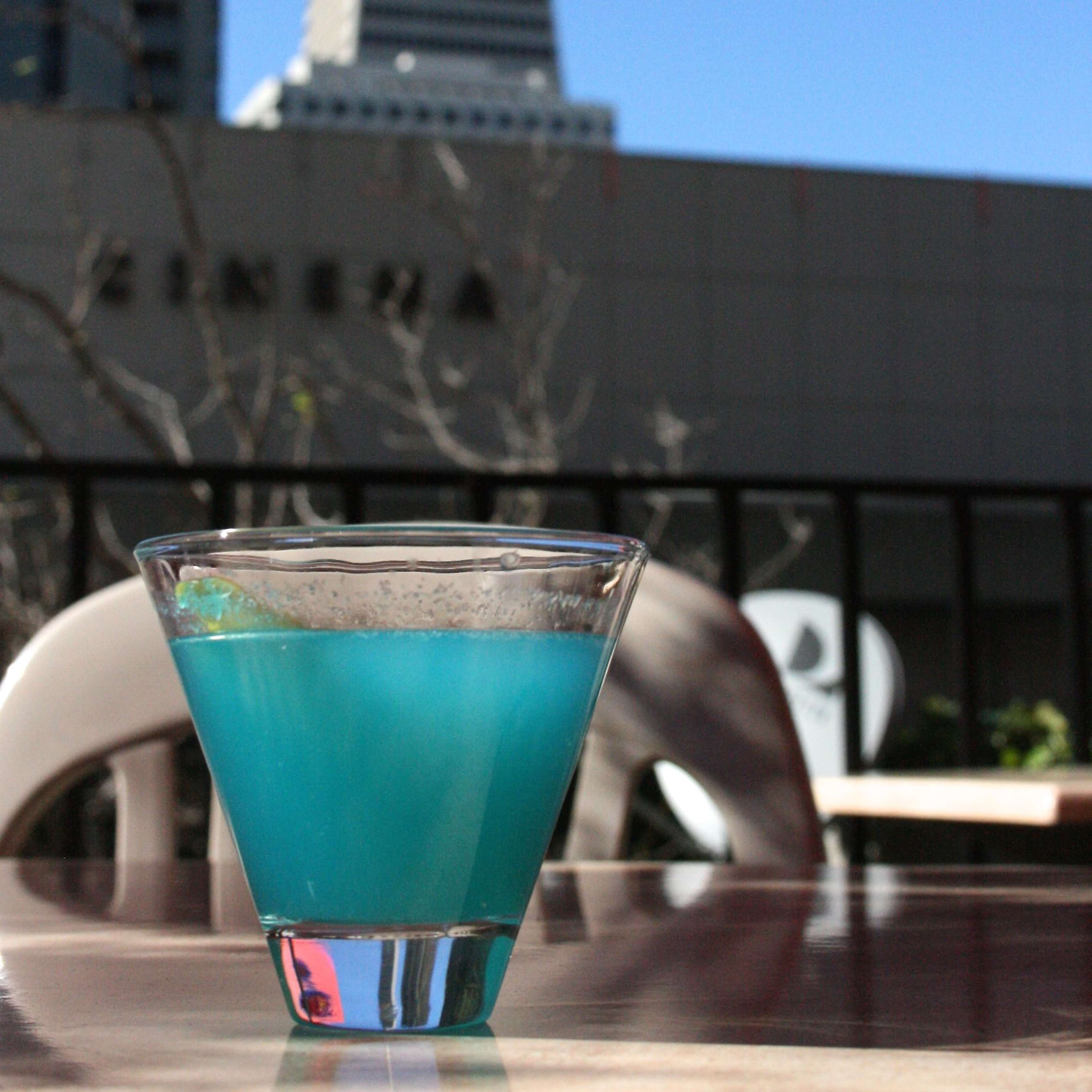 A blue cocktail is placed in a glass on a table with a high-rise building and a tower in the background under a clear blue sky.