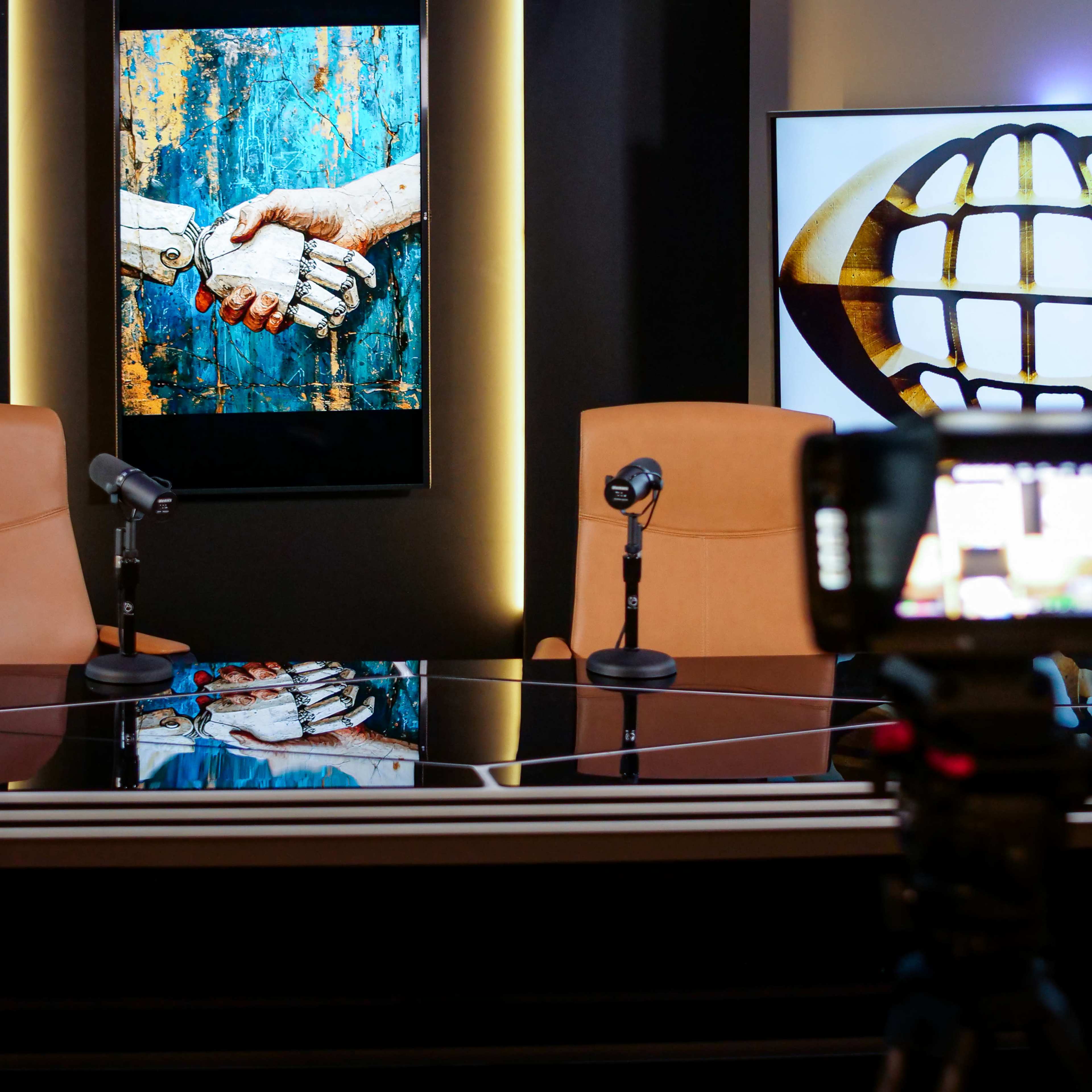 A modern studio setting with two empty chairs at a table, flanked by large screens displaying an artwork of a robotic handshake and a globe symbol.
