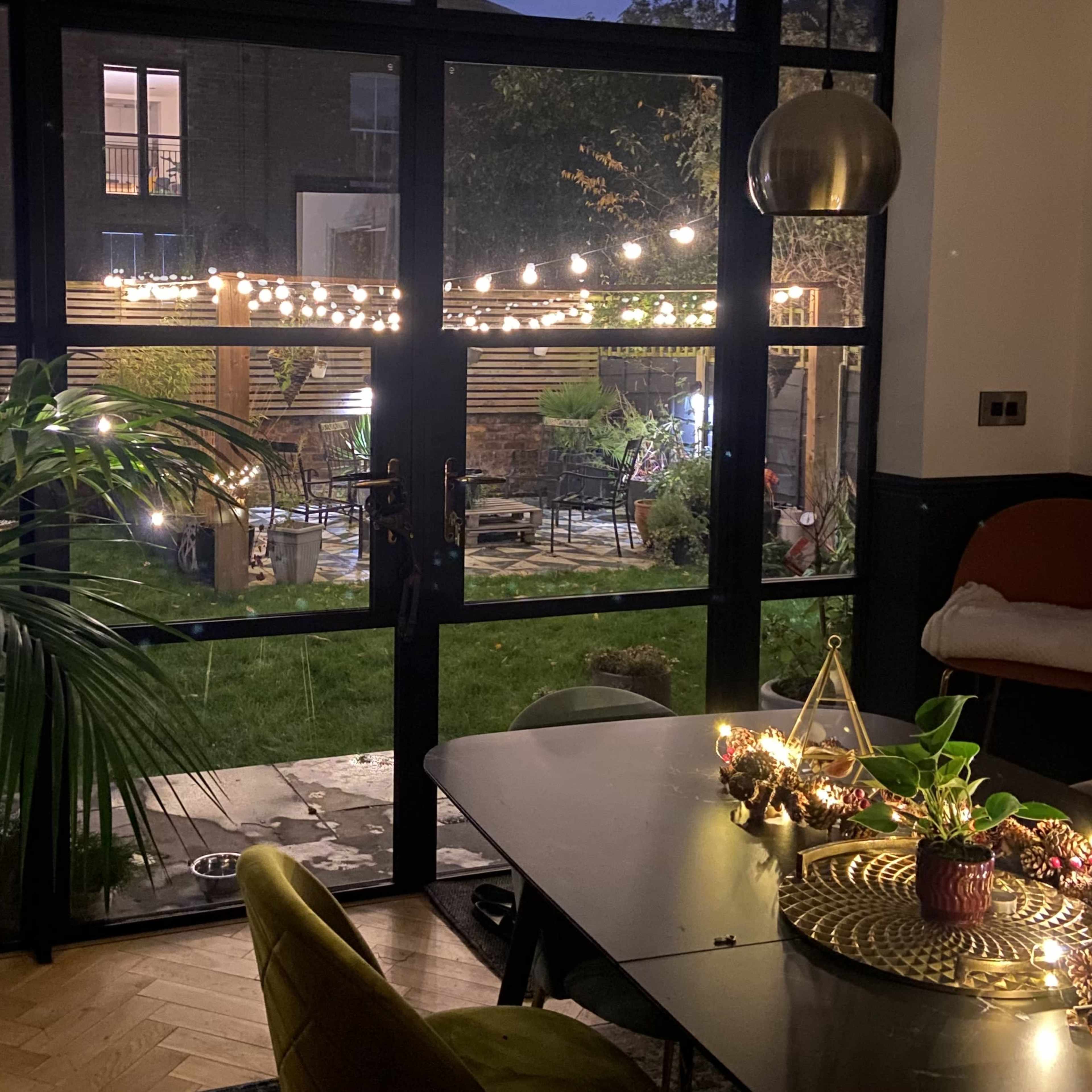 A cozy dining area overlooks a well-lit garden through large glass doors adorned with string lights.