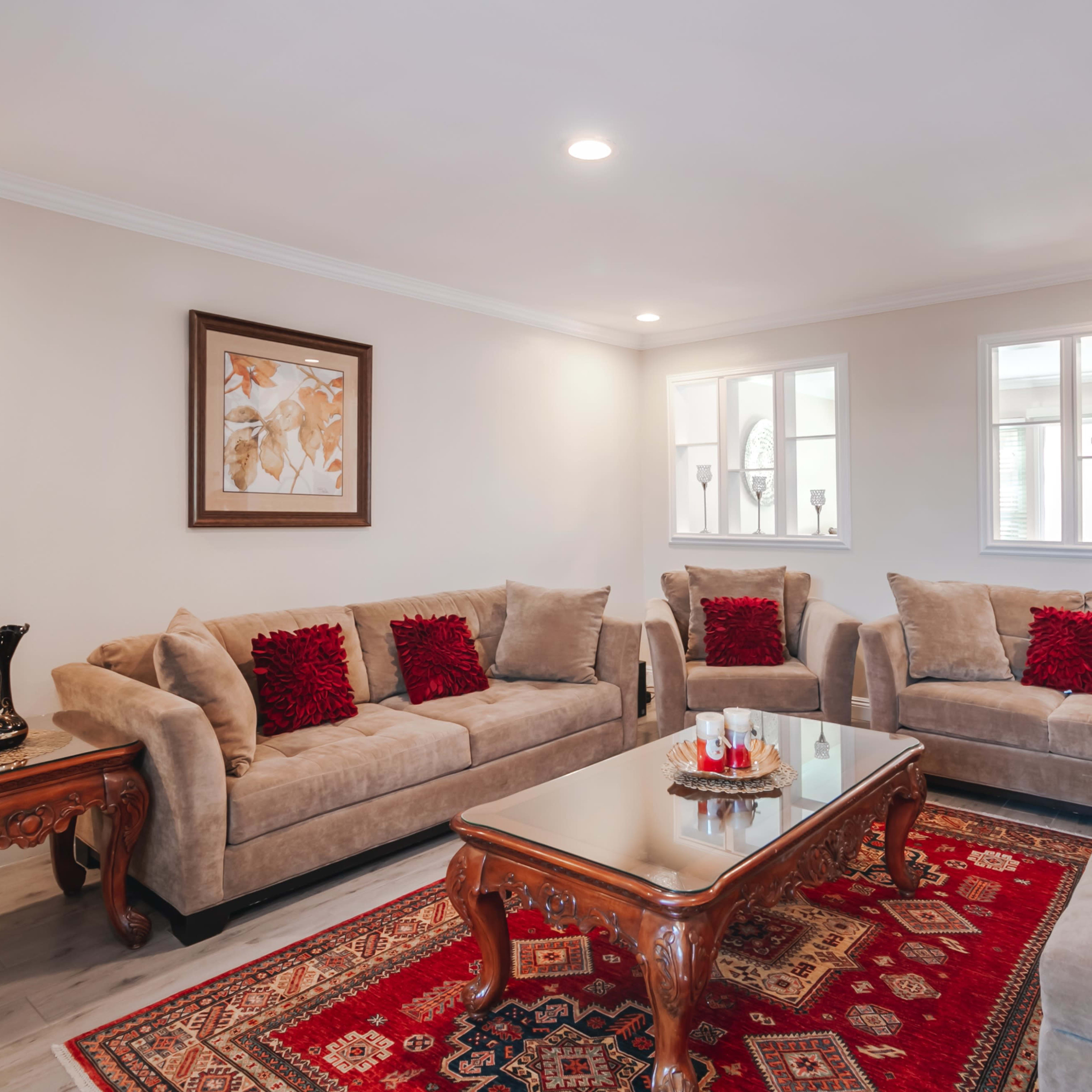 A living room features two sofas and two armchairs arranged around a coffee table on a patterned rug, with decorative pillows and a framed picture on the wall.