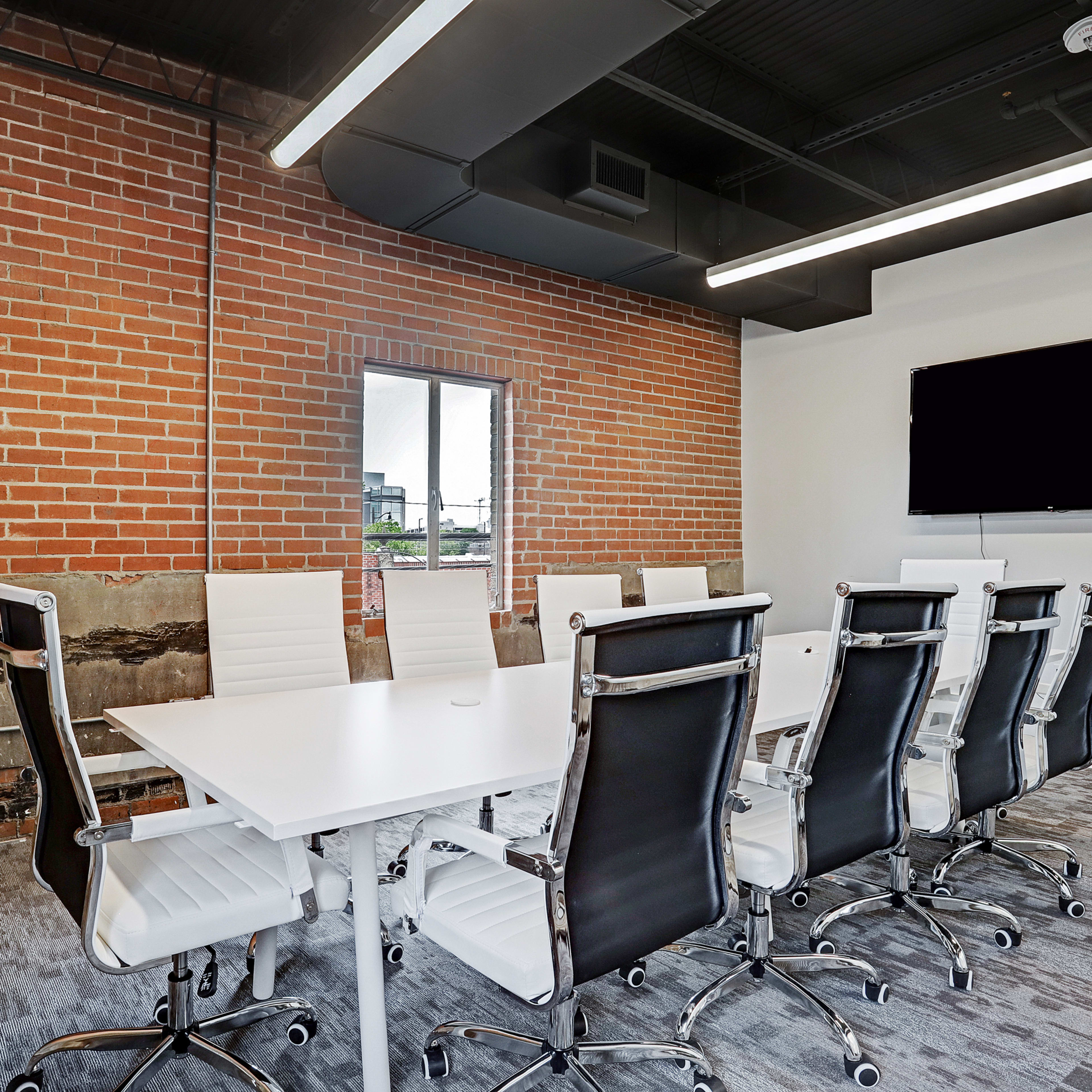 A modern conference room features a long white table surrounded by twelve black ergonomic chairs, with exposed brick walls and a large screen mounted on the opposite wall.