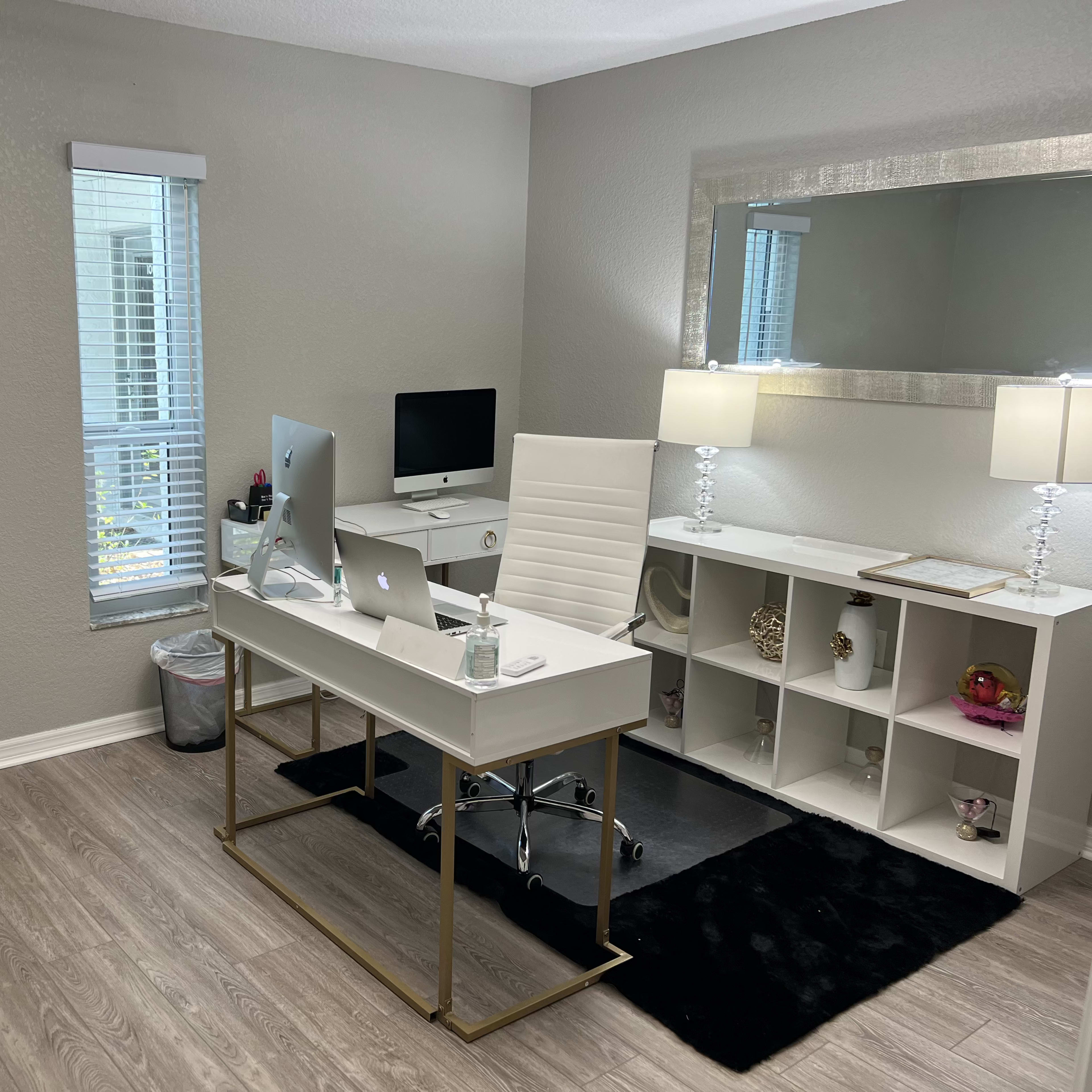 A modern office space features a white desk with a computer, a white ergonomic chair, a matching decorative table with lamps, and a storage unit against a light-colored wall.